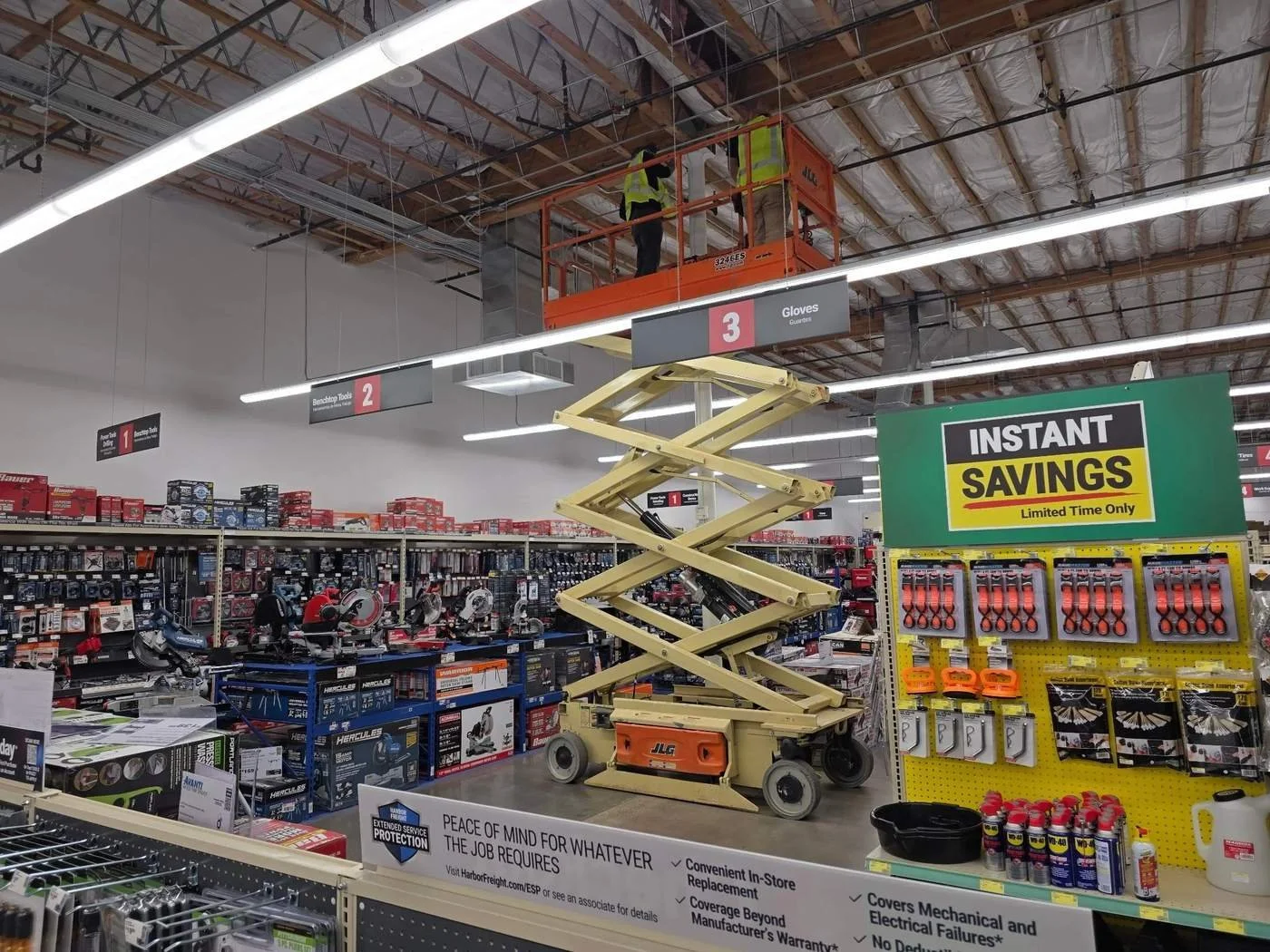 Two workers in safety vests working on ceiling panels on a lift inside a hardware store, with shelves of tools and a sign advertising instant savings.