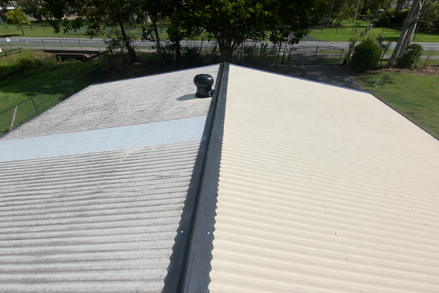 View of a roof with two sections, one dirty and the other clean, separated by a ridge with a black vent on top, surrounded by trees, a fence, and a road in the background.