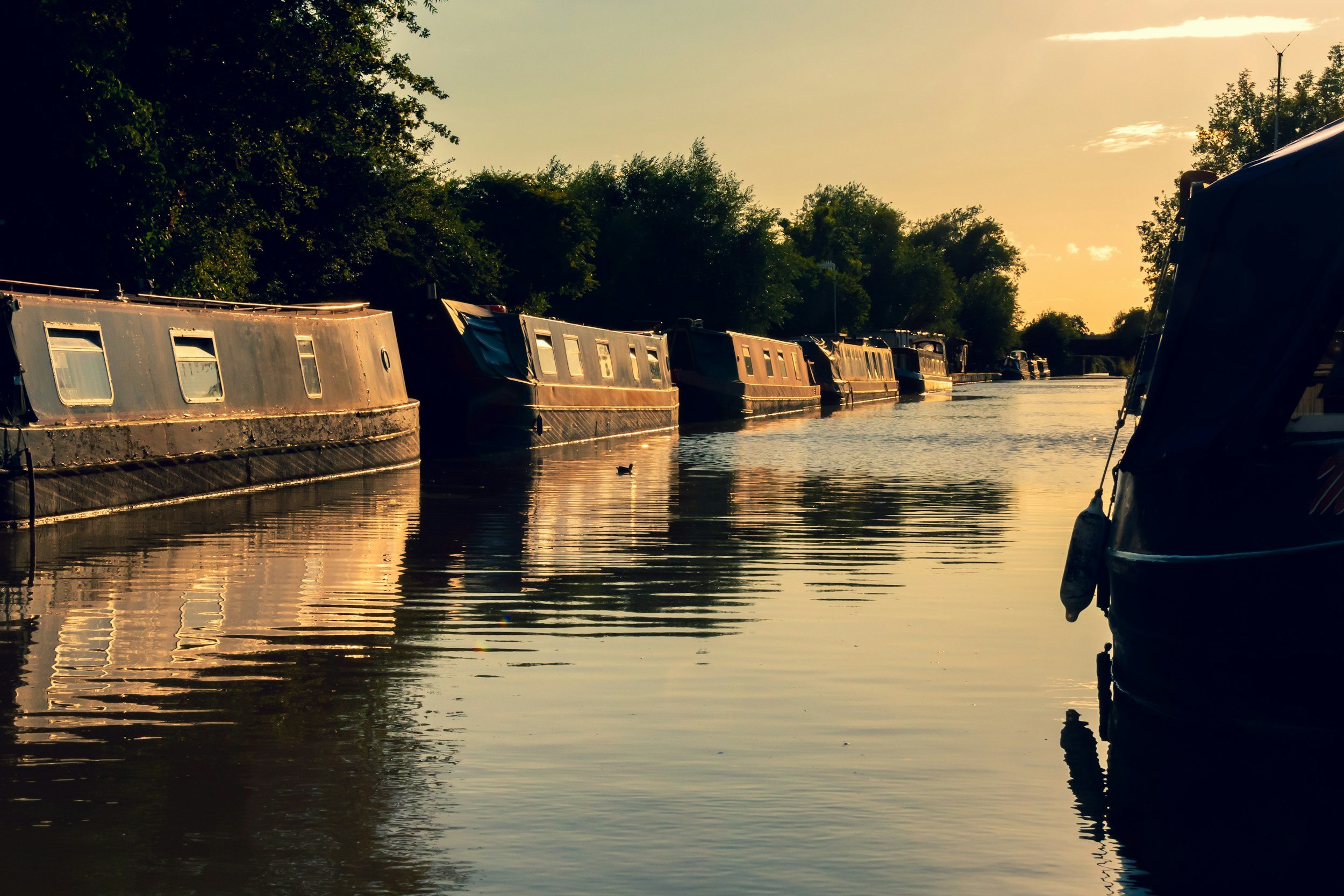 Boats docked along a peaceful river at sunset, with trees lining the riverbank and the sky casting a warm glow.
