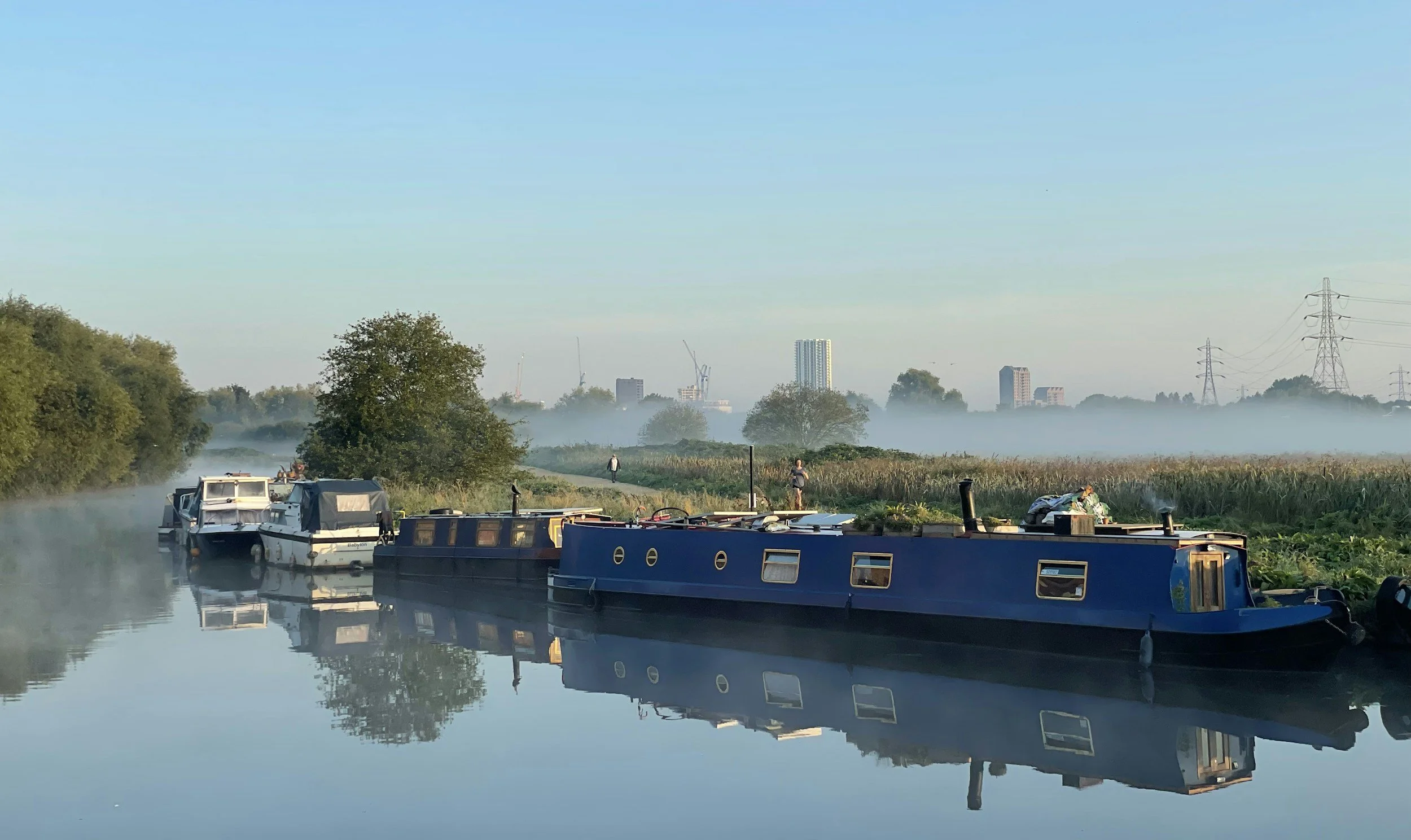 Boats moored on a calm river with fog rising, greenery on the banks, a few people walking, and city buildings with cranes in the background under a clear blue sky.
