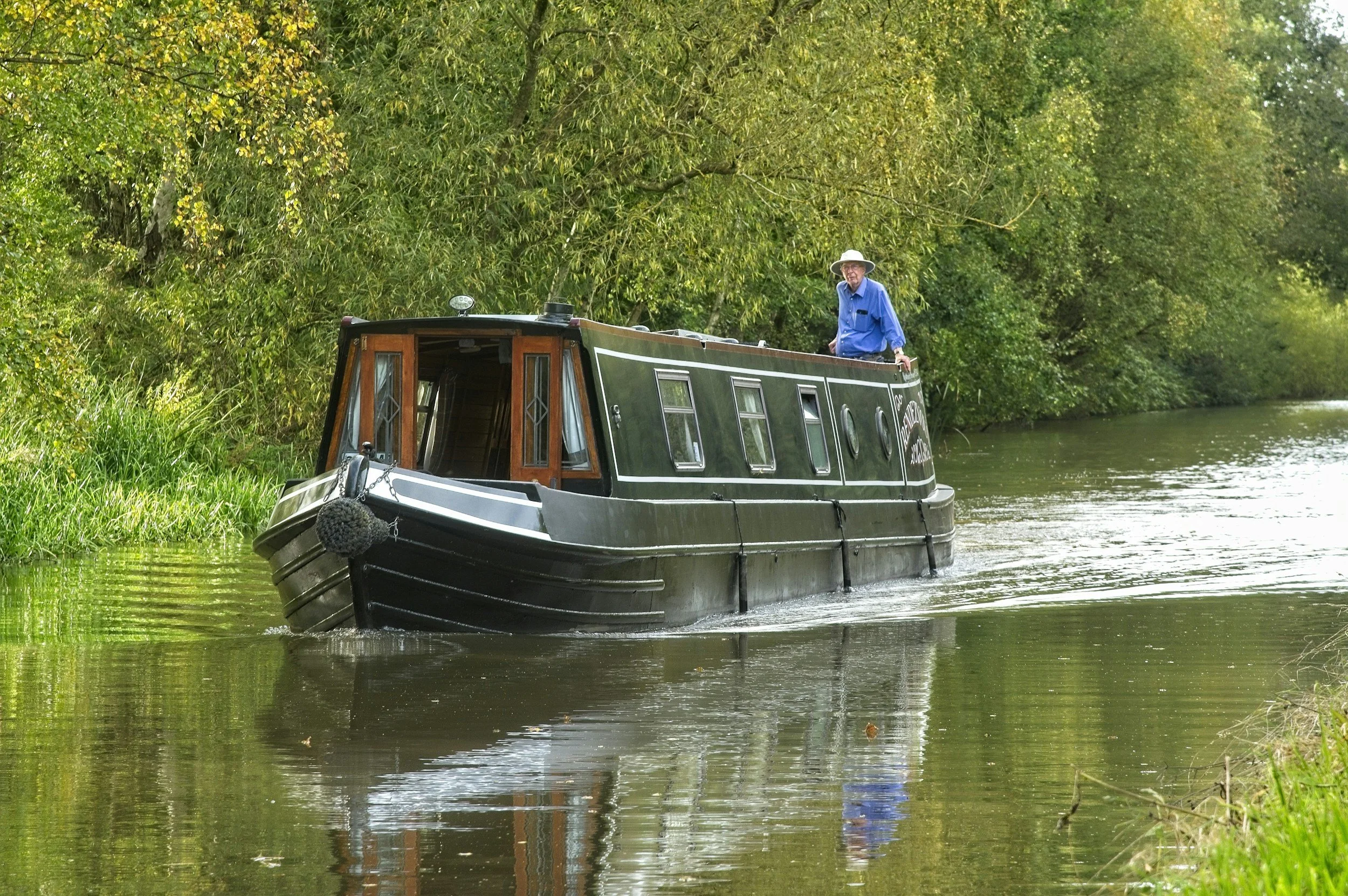 A man with a hat in a blue jacket standing at the open roof of a black narrowboat moving through a green, wooded waterway.