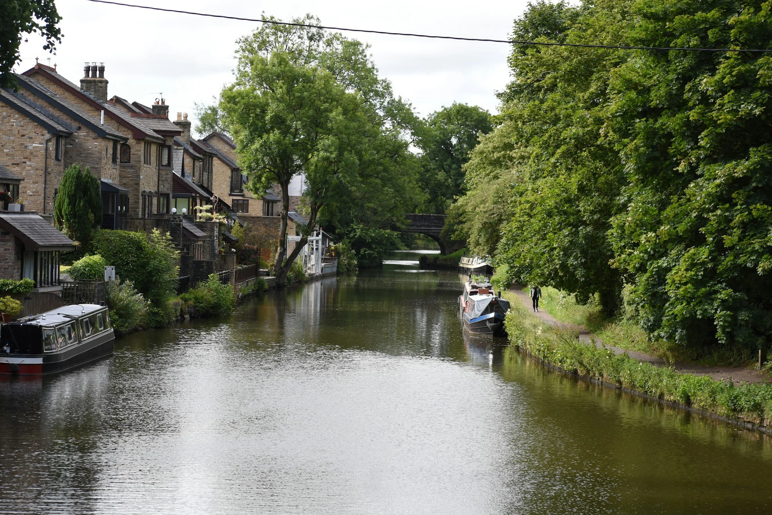A peaceful canal lined with houseboats and trees, with a person walking along the path on the right side.