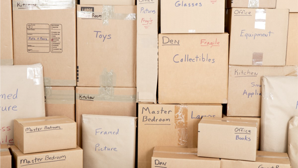 Stacked cardboard boxes labeled with room names and contents in a storage area.
