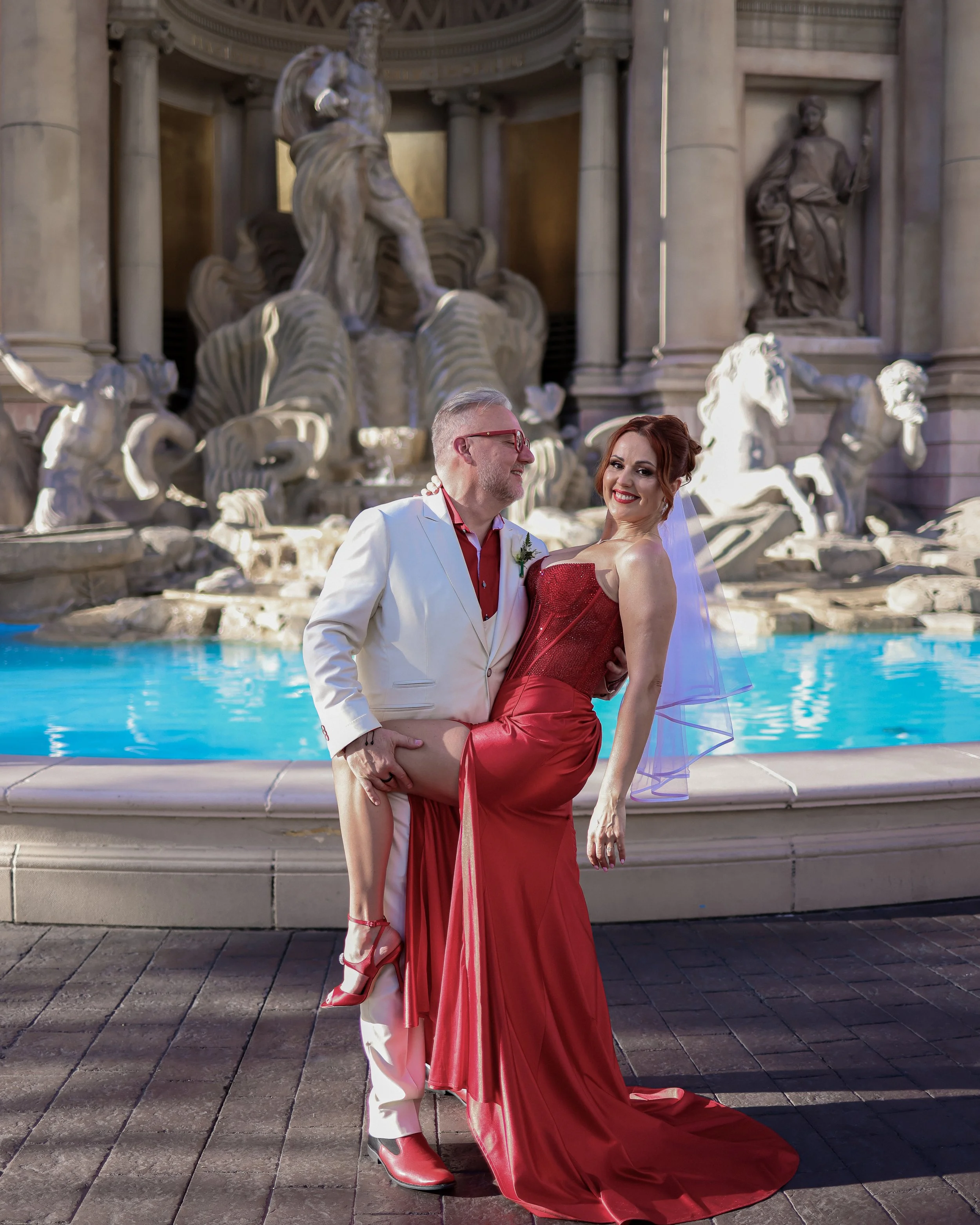 A couple dressed in formal wedding attire standing in front of a large ornate fountain with multiple sculptures, including horses and figures, on a paved area outside a historic building.