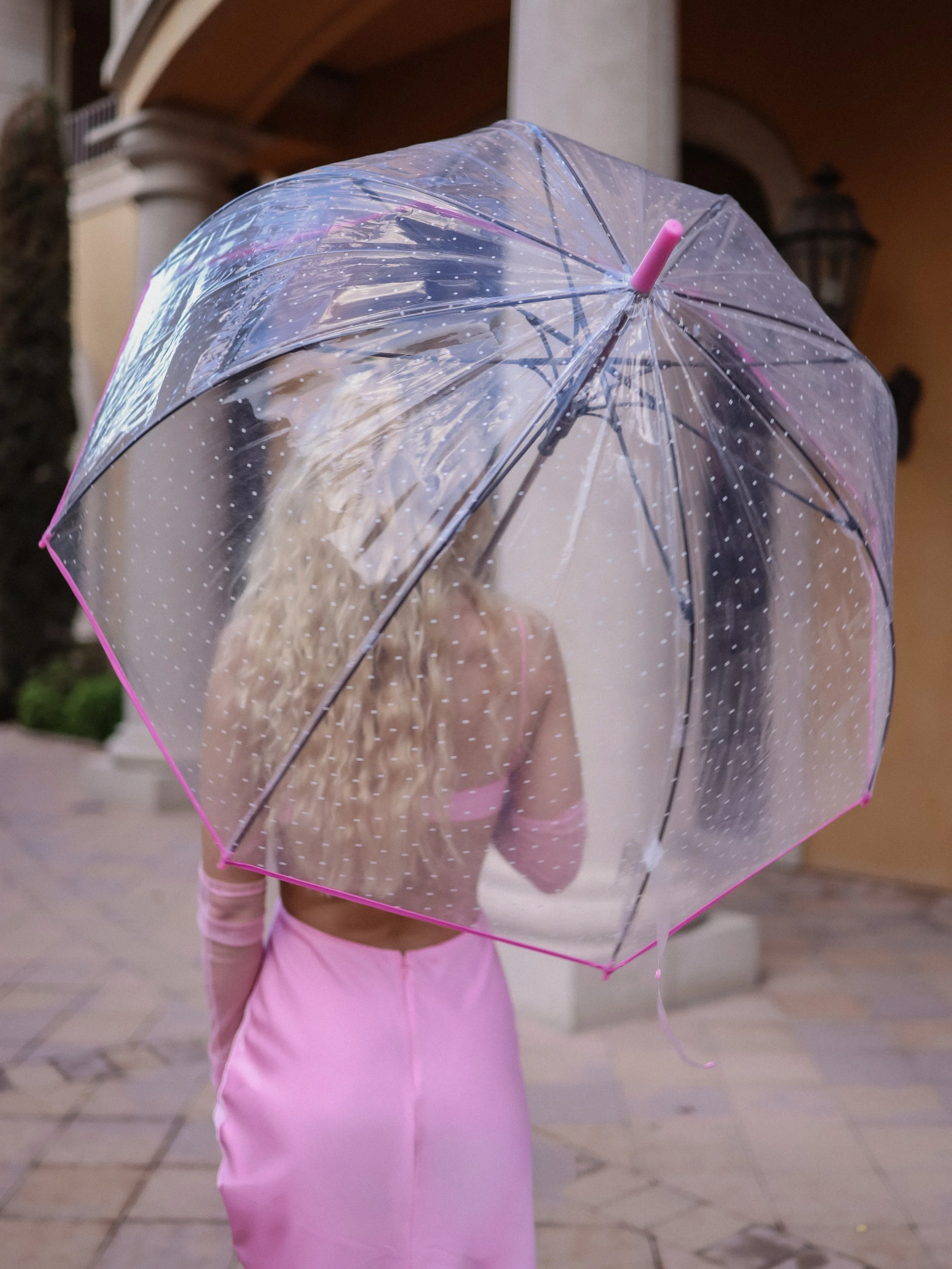 Child holding a transparent umbrella with white polka dots, wearing pink pants and standing outdoors.