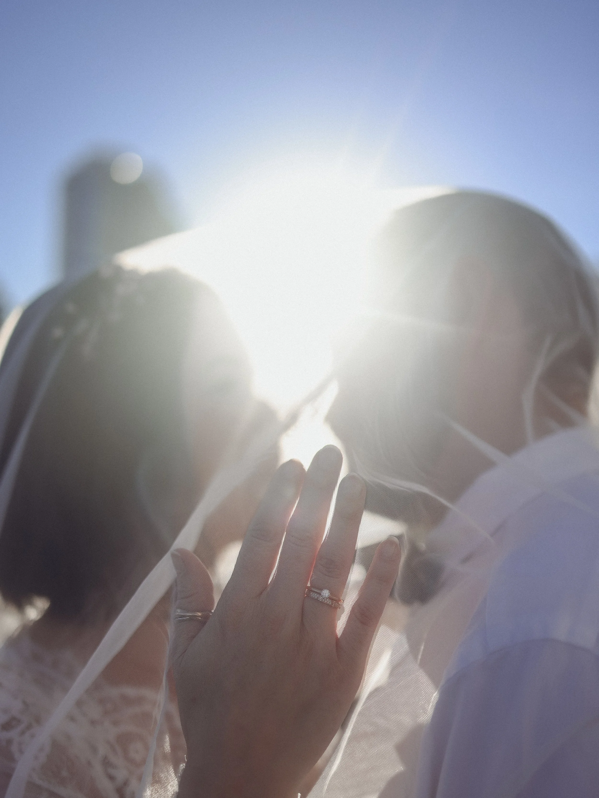 Close-up view of a person's hand with rings, pressing or touching a sheer white fabric or curtain, sunlight illuminating through, with a clear blue sky in the background.