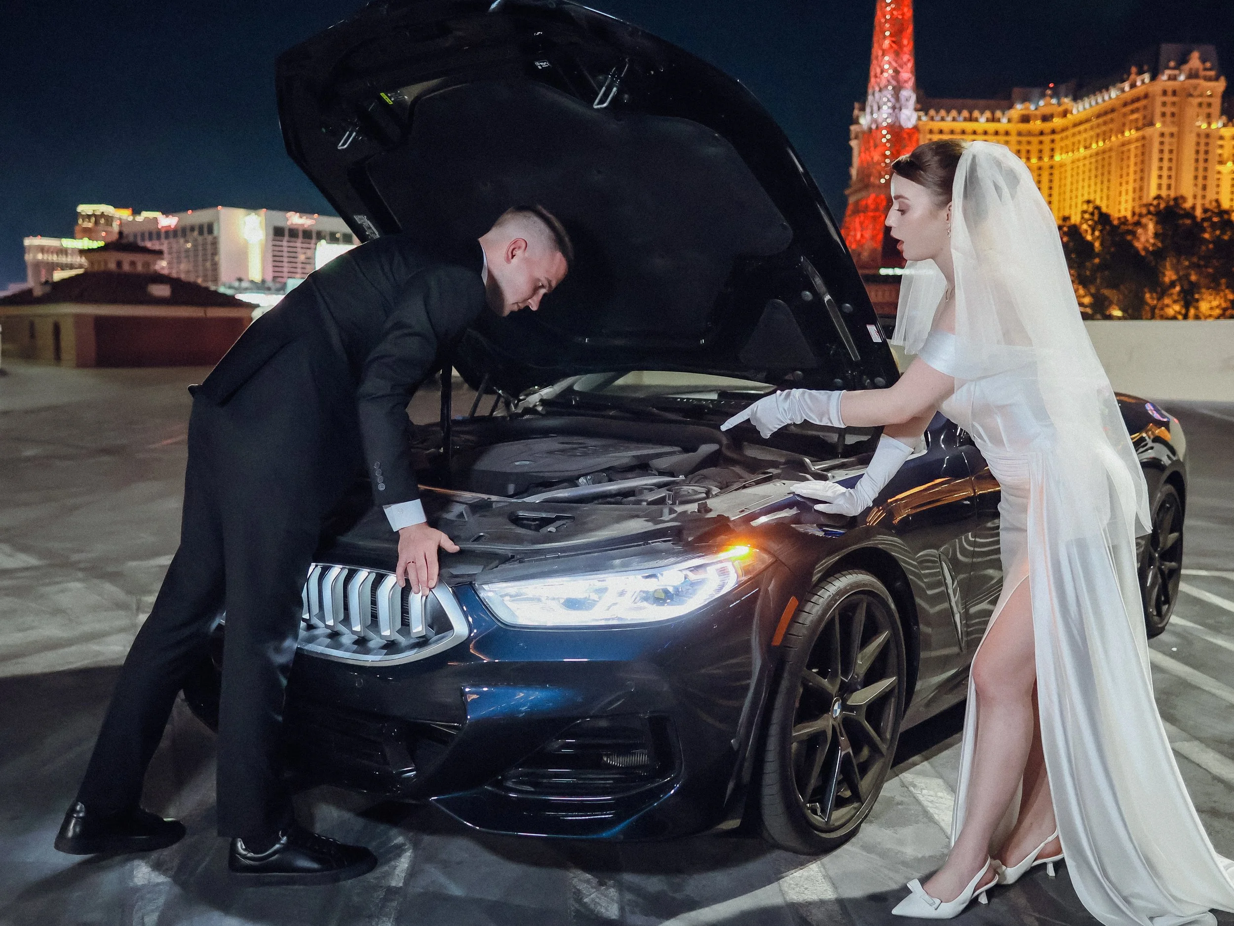 A person in a black suit, likely a man, is leaning into the open hood of a black luxury car, possibly an SUV, on a rooftop parking structure at night. A woman in a bridal dress with a veil is standing next to the car, pointing into the engine area. T