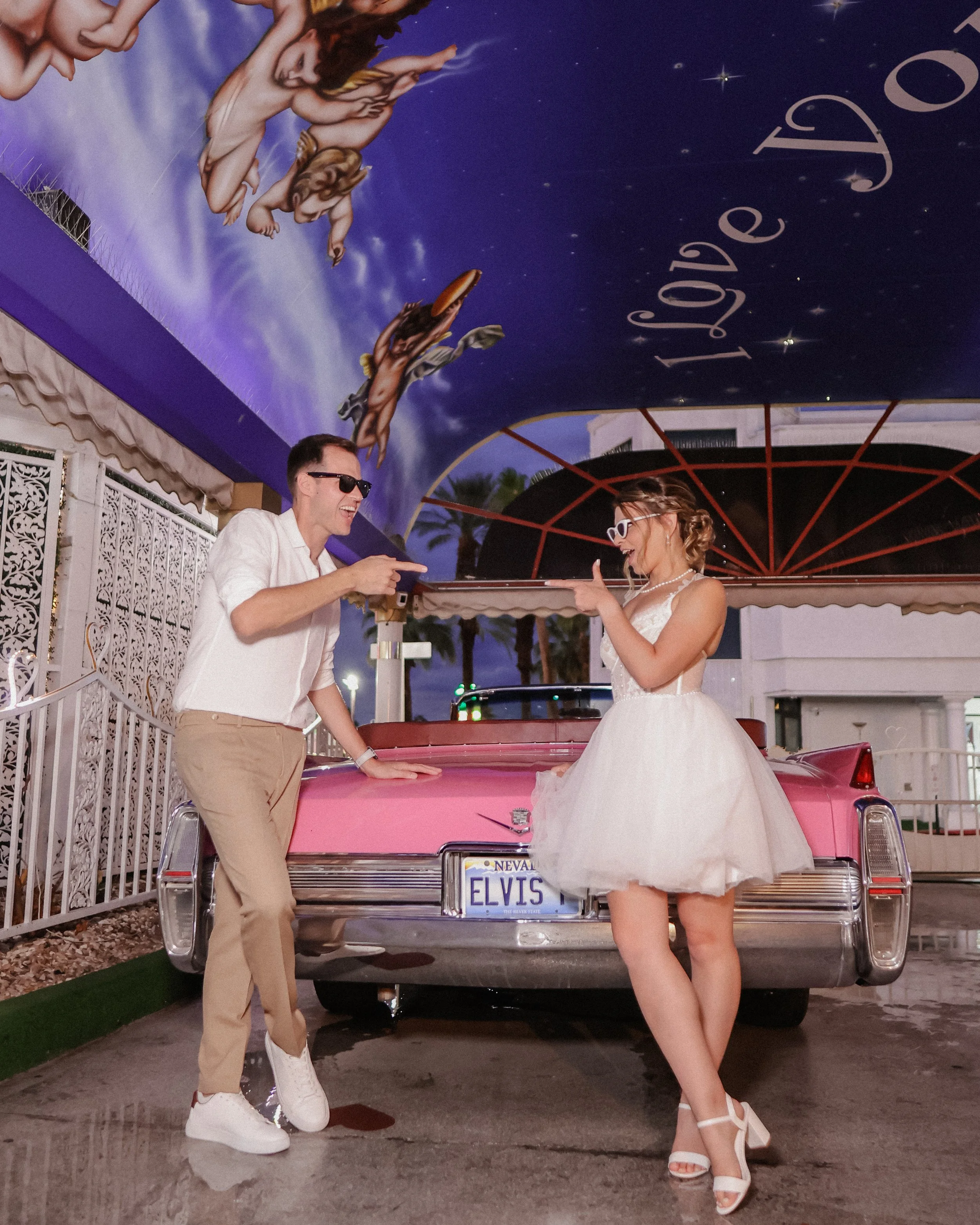 A man and woman wearing sunglasses and vintage-style outfits posing in front of a pink Cadillac with a Reno Nevada license plate reading Elvis. They are under a colorful, painted canopy with painted angels and the words "Love D" visible.
