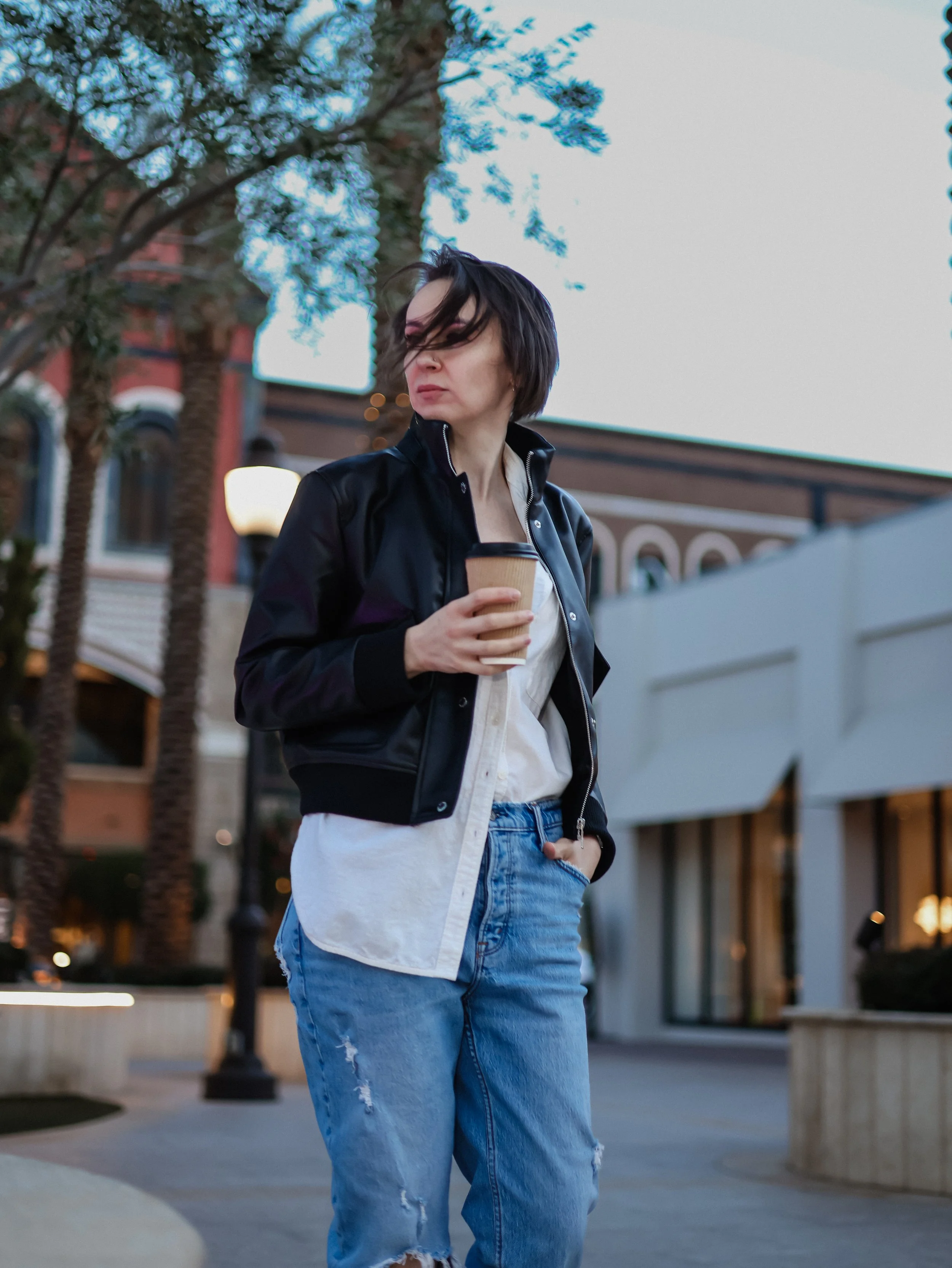 A woman with black hair and a nose piercing holding a disposable coffee cup in an outdoor shopping area during early evening, wearing a black jacket and ripped blue jeans.