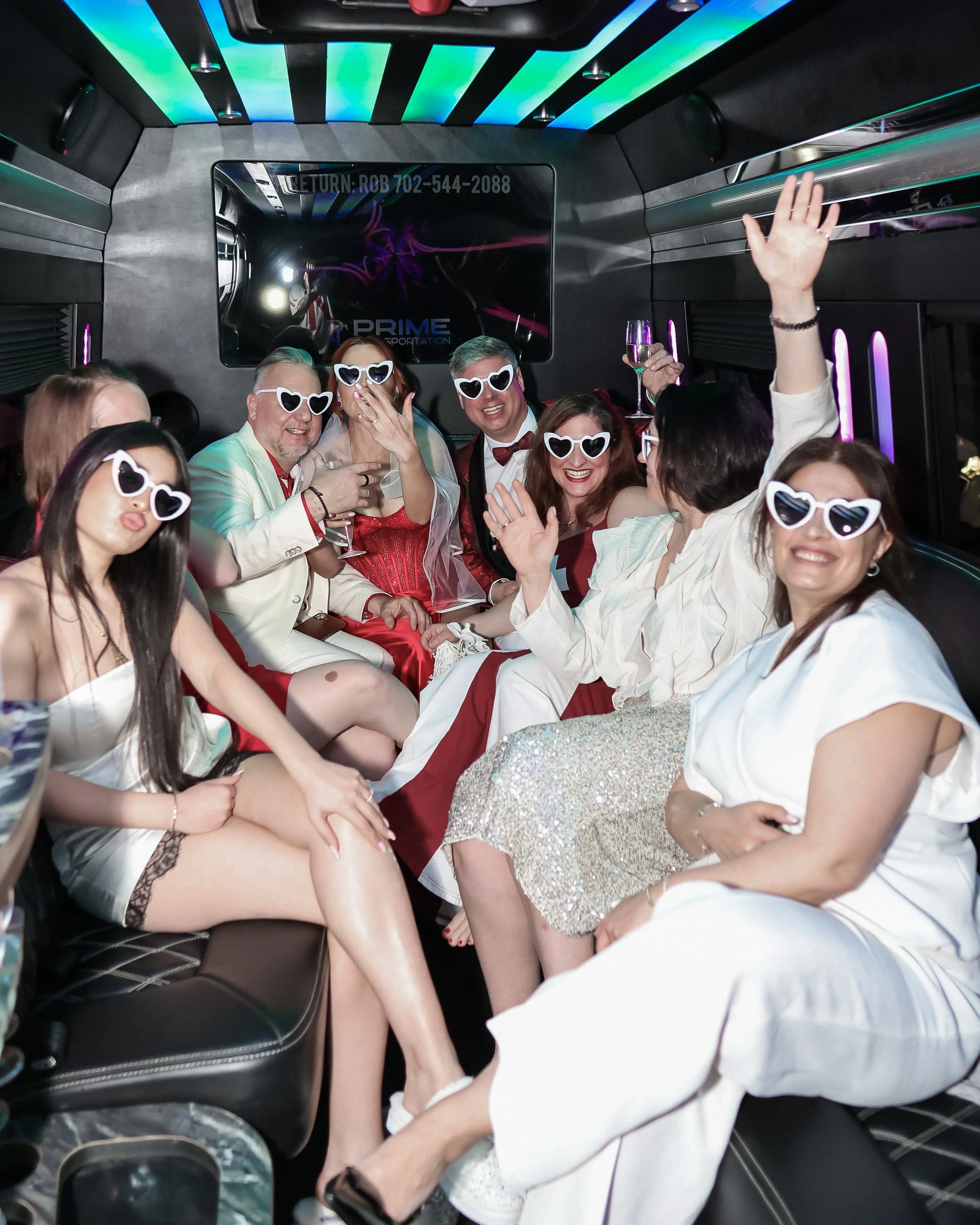 Group of people dressed in formal attire inside a limousine with colorful lighting, celebrating and smiling, all wearing heart-shaped sunglasses.