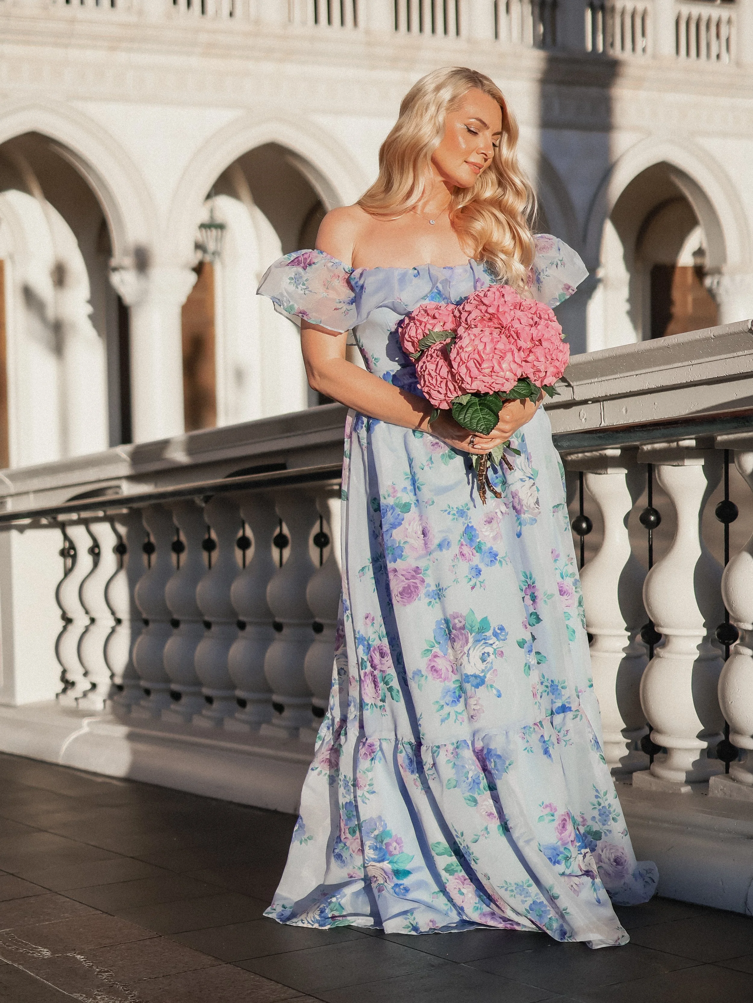 A woman in a long floral dress holding a bouquet of pink hydrangeas on a balcony with arches.