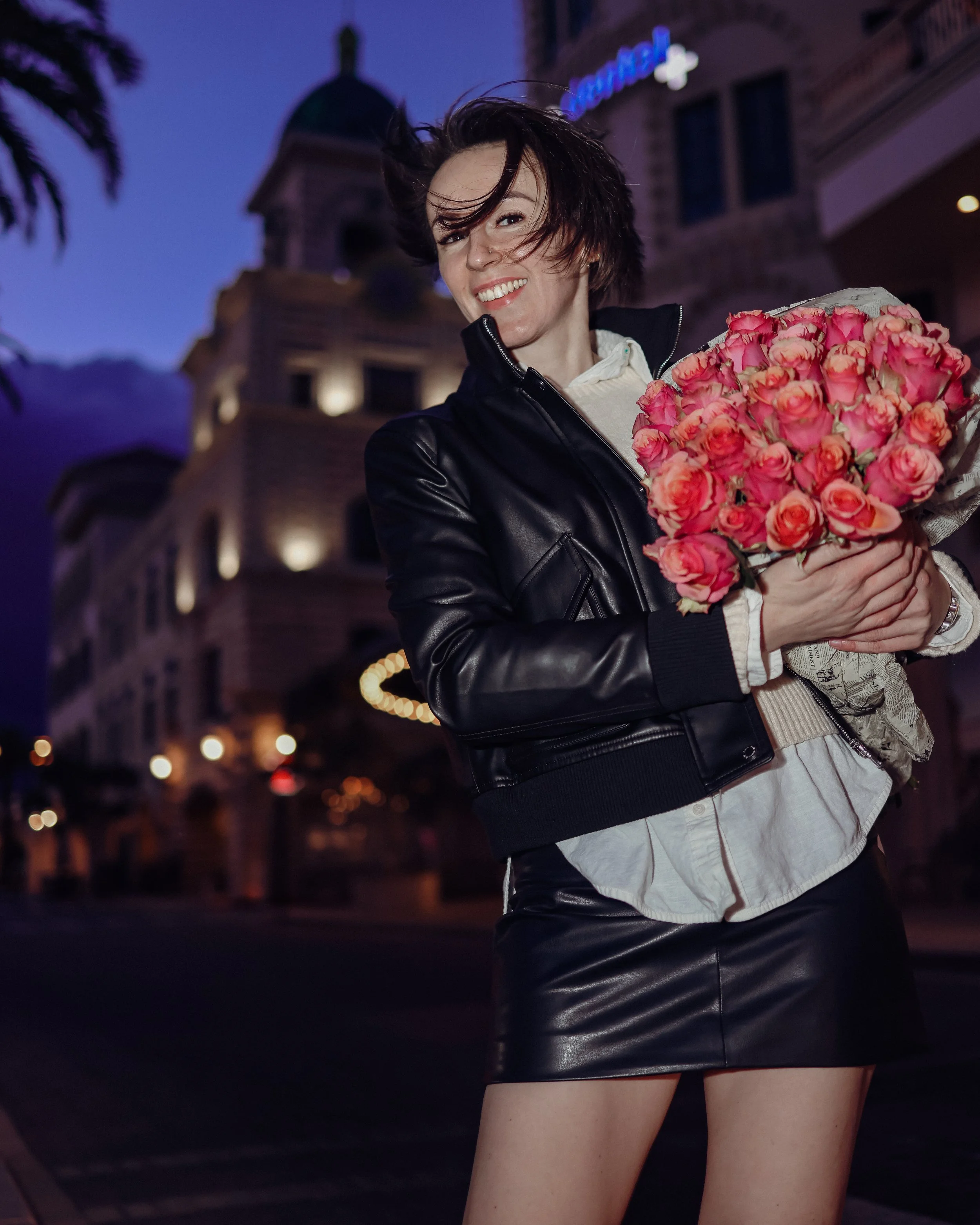 A woman with wind-blown hair holding a large bouquet of pink roses on a city street at dusk, with a historic building and purple sky in the background.