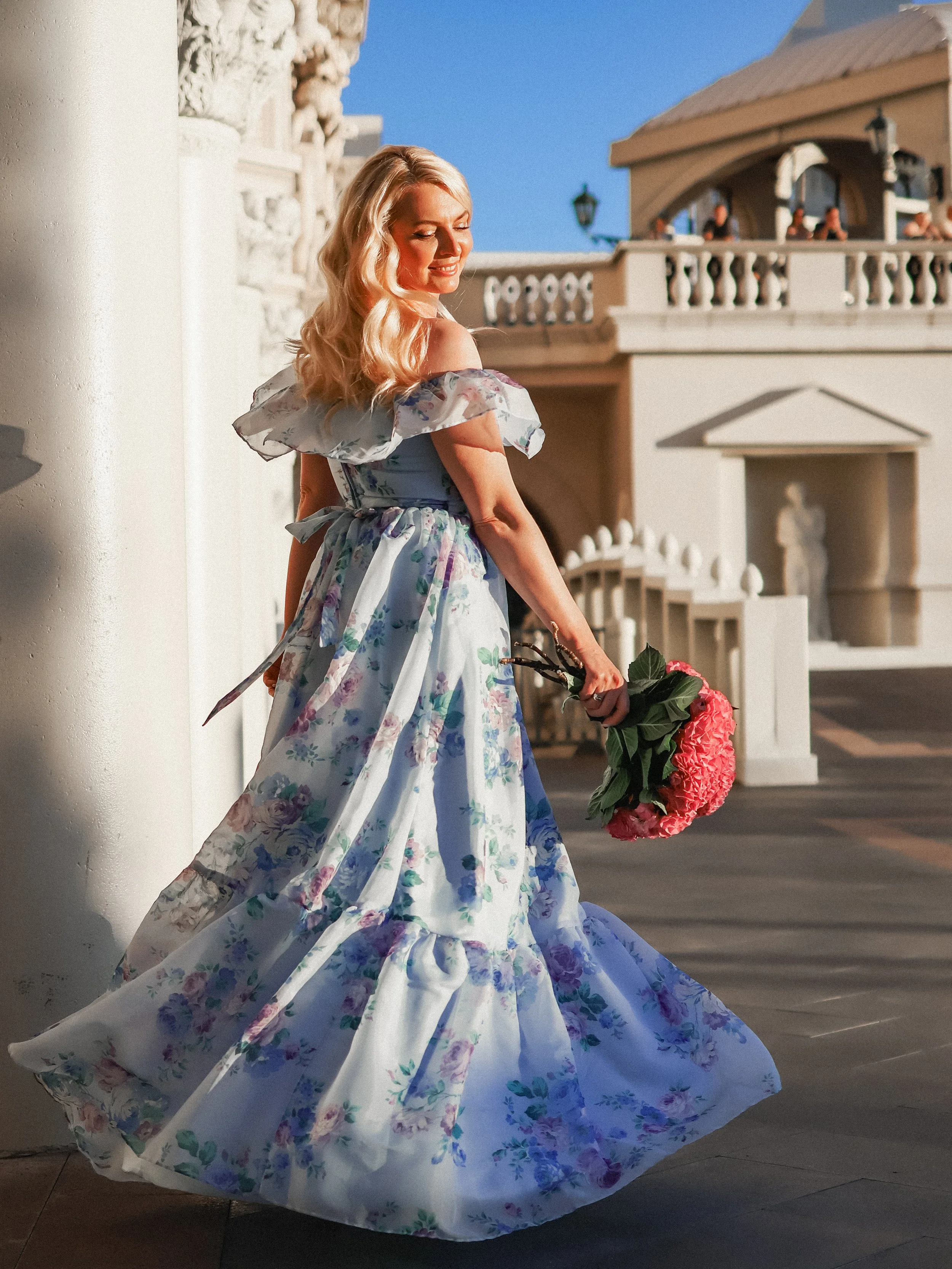 A woman with long blonde hair wearing a floral, off-the-shoulder gown, holding a bouquet of pink flowers, standing outdoors near a white column and building with a balcony, under a blue sky.