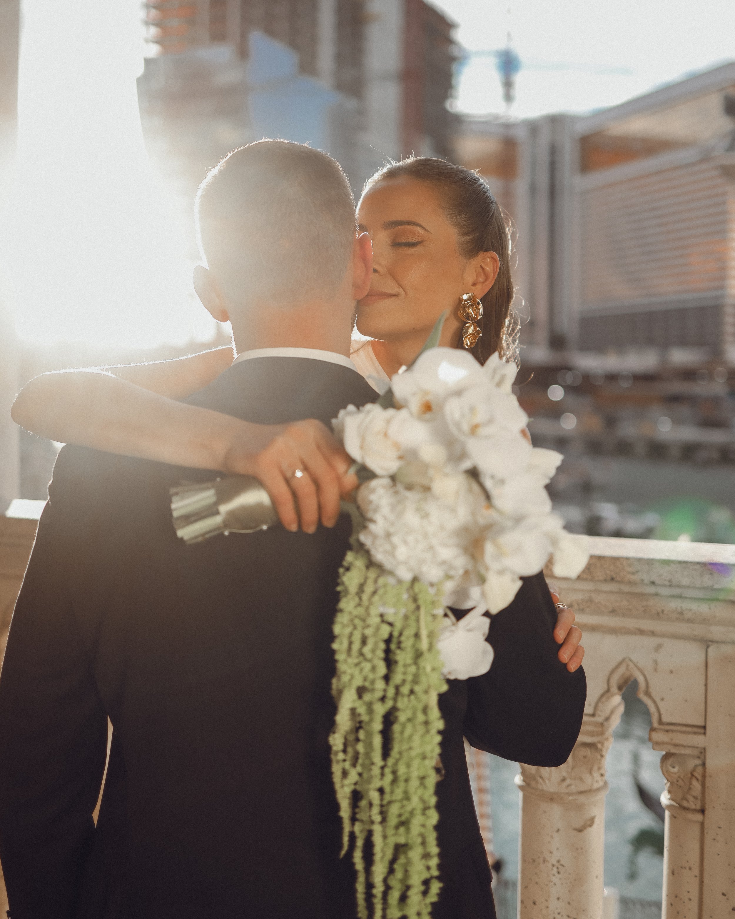 A bride and groom embrace on a balcony with sunlight behind them, with city buildings in the background. The bride is holding a white floral bouquet, and both are dressed in formal wedding attire.