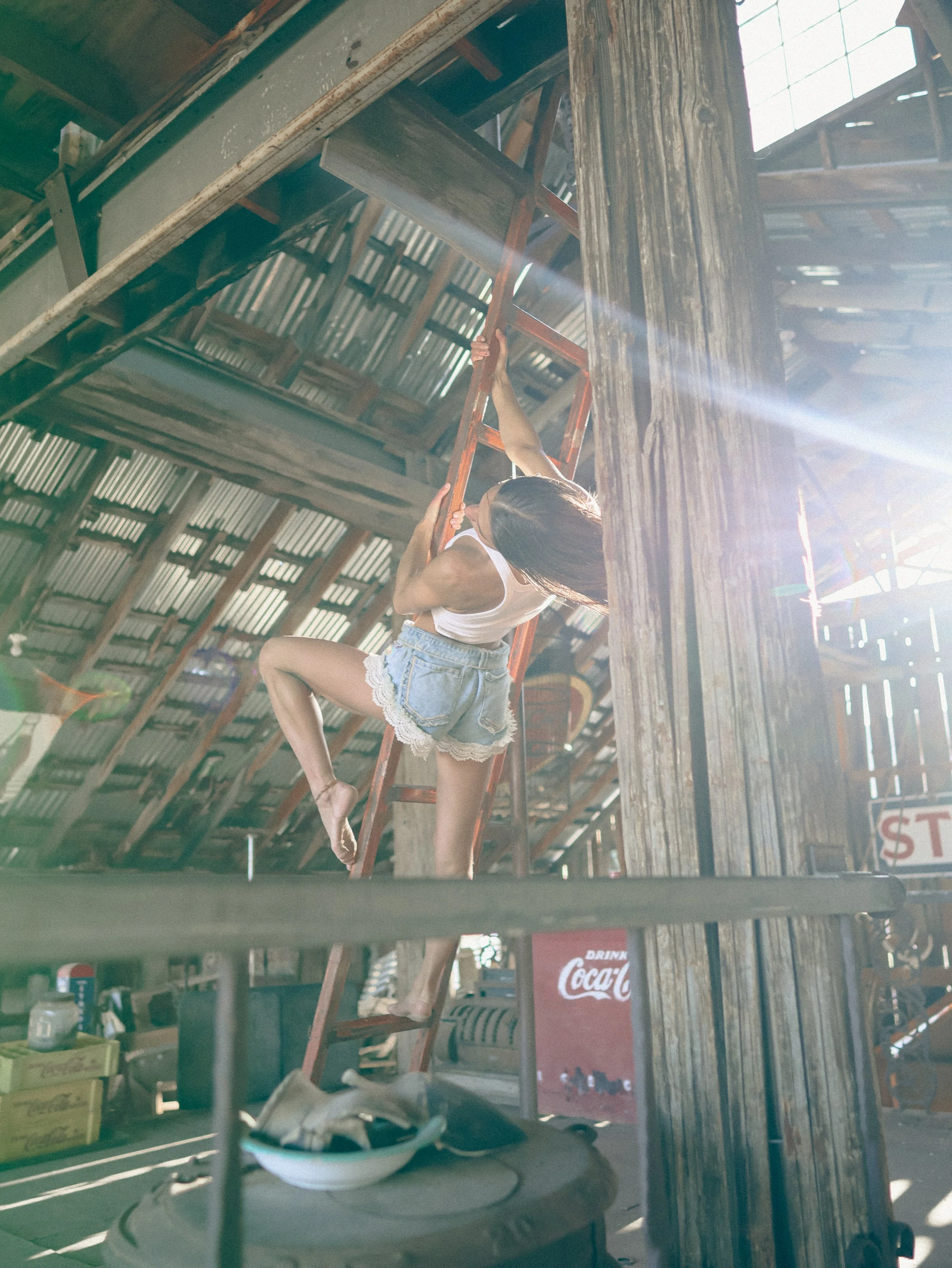 A person climbing an orange metal ladder inside a rustic wooden barn, with sunlight streaming through the roof, and various snacks and drinks on a table in the foreground.
