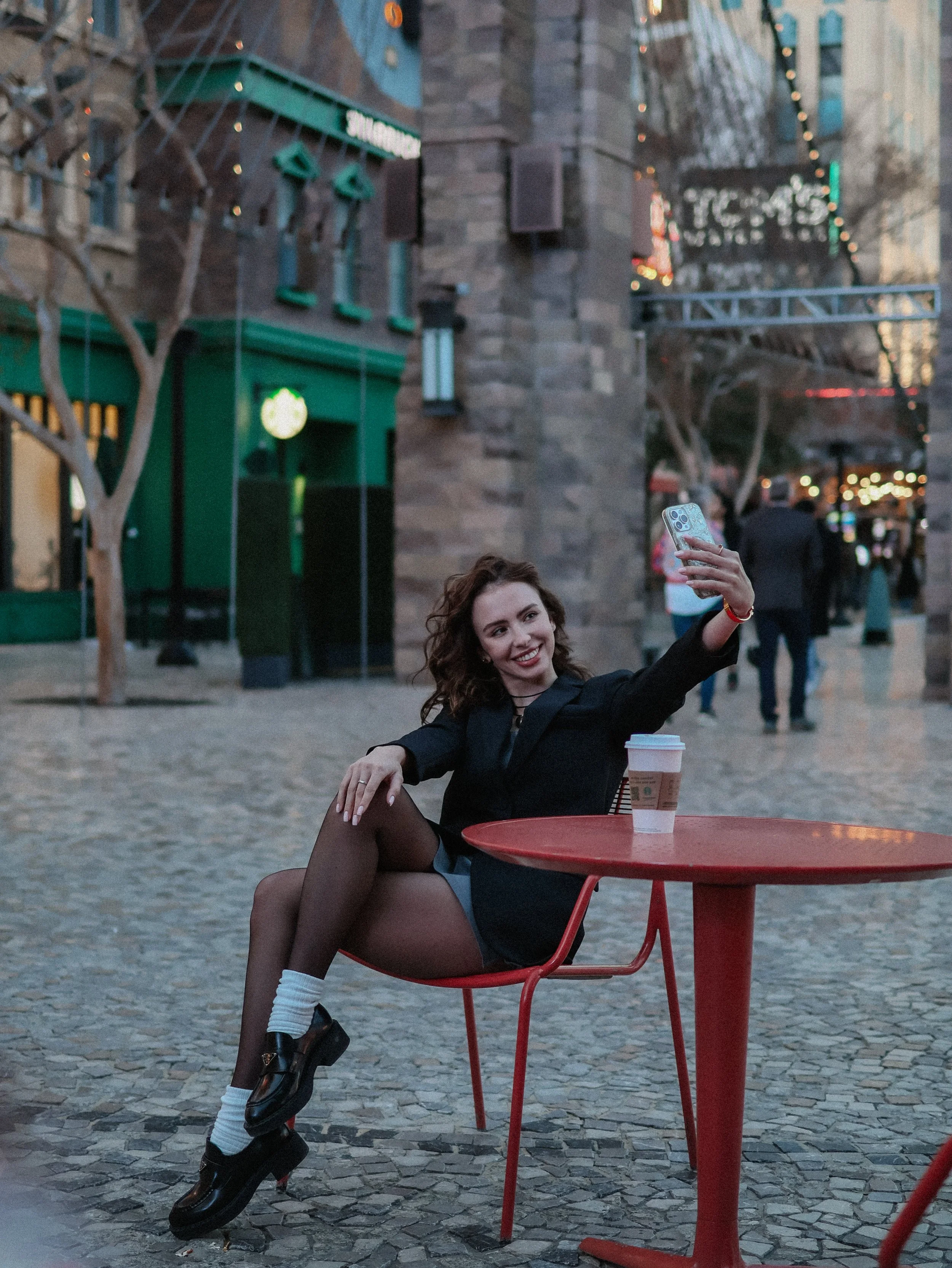 A young woman with curly hair dressed in a black outfit and black lace-up shoes sitting at a red outdoor table, smiling and taking a selfie with her phone, with a coffee cup on the table, in a city square during evening with festive lights and buildi