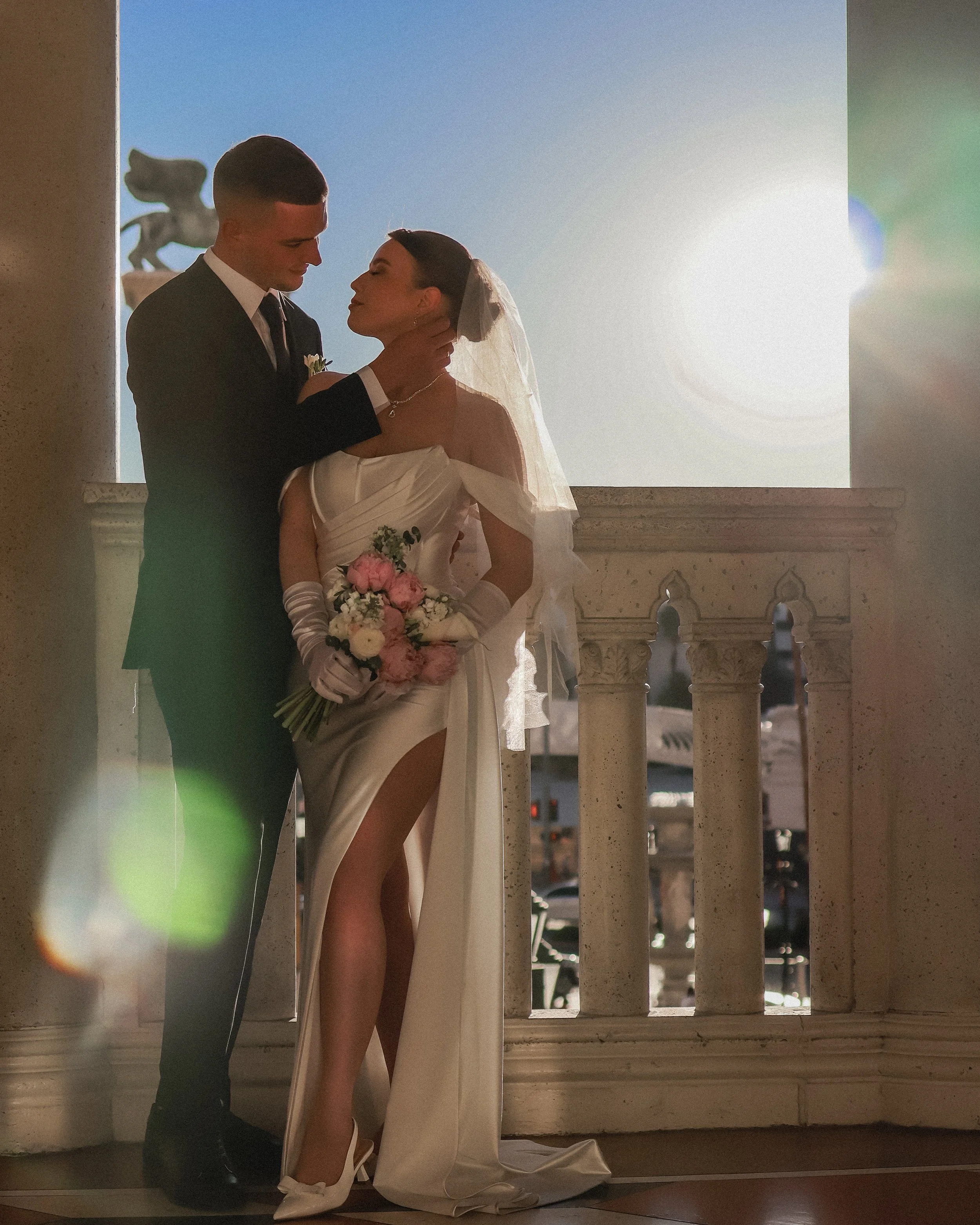 A bride and groom standing close inside a building on a balcony, with a castle-like structure and a lion statue in the background, sharing an intimate moment.