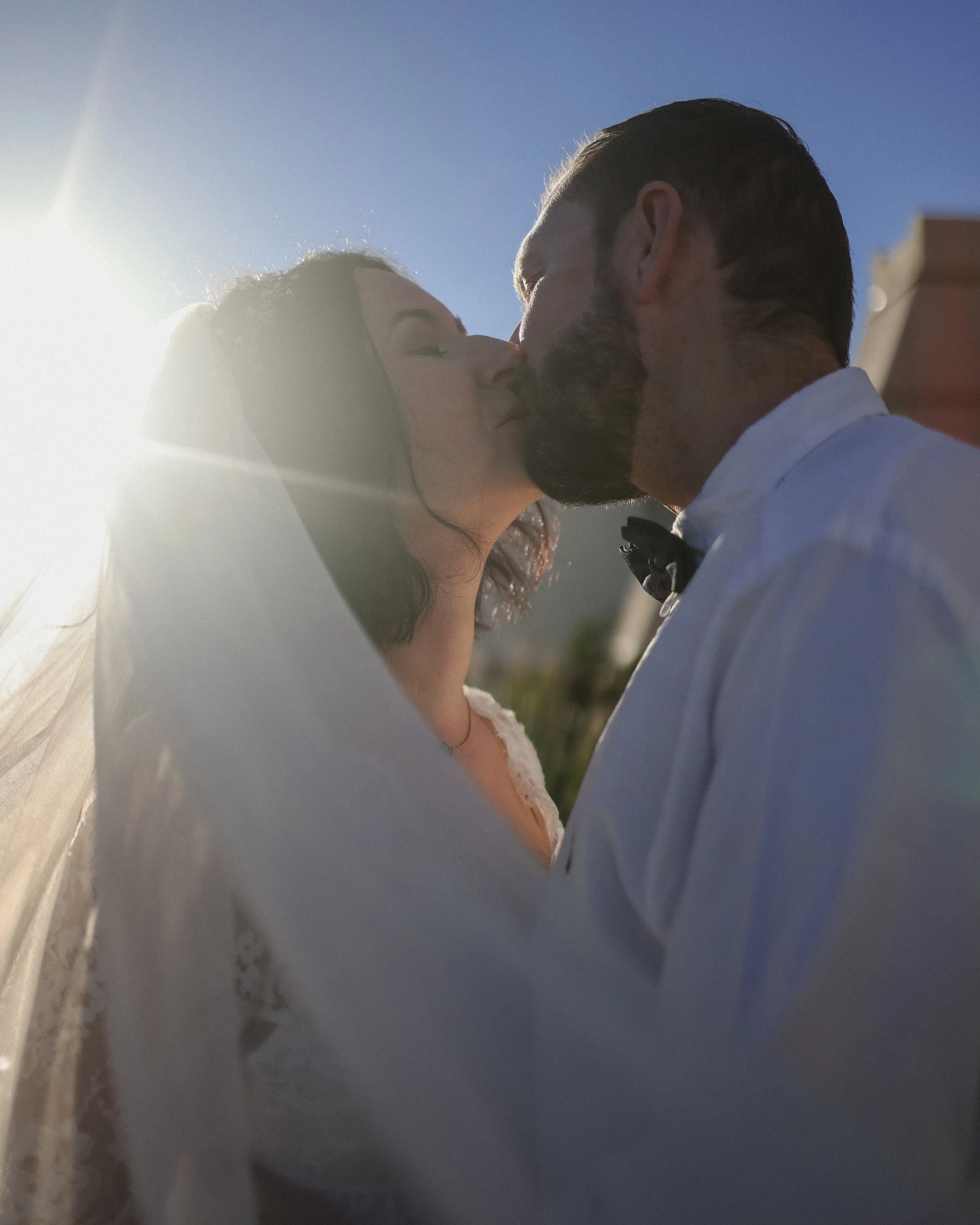 A couple kissing outside during daytime with a blue sky behind them.
