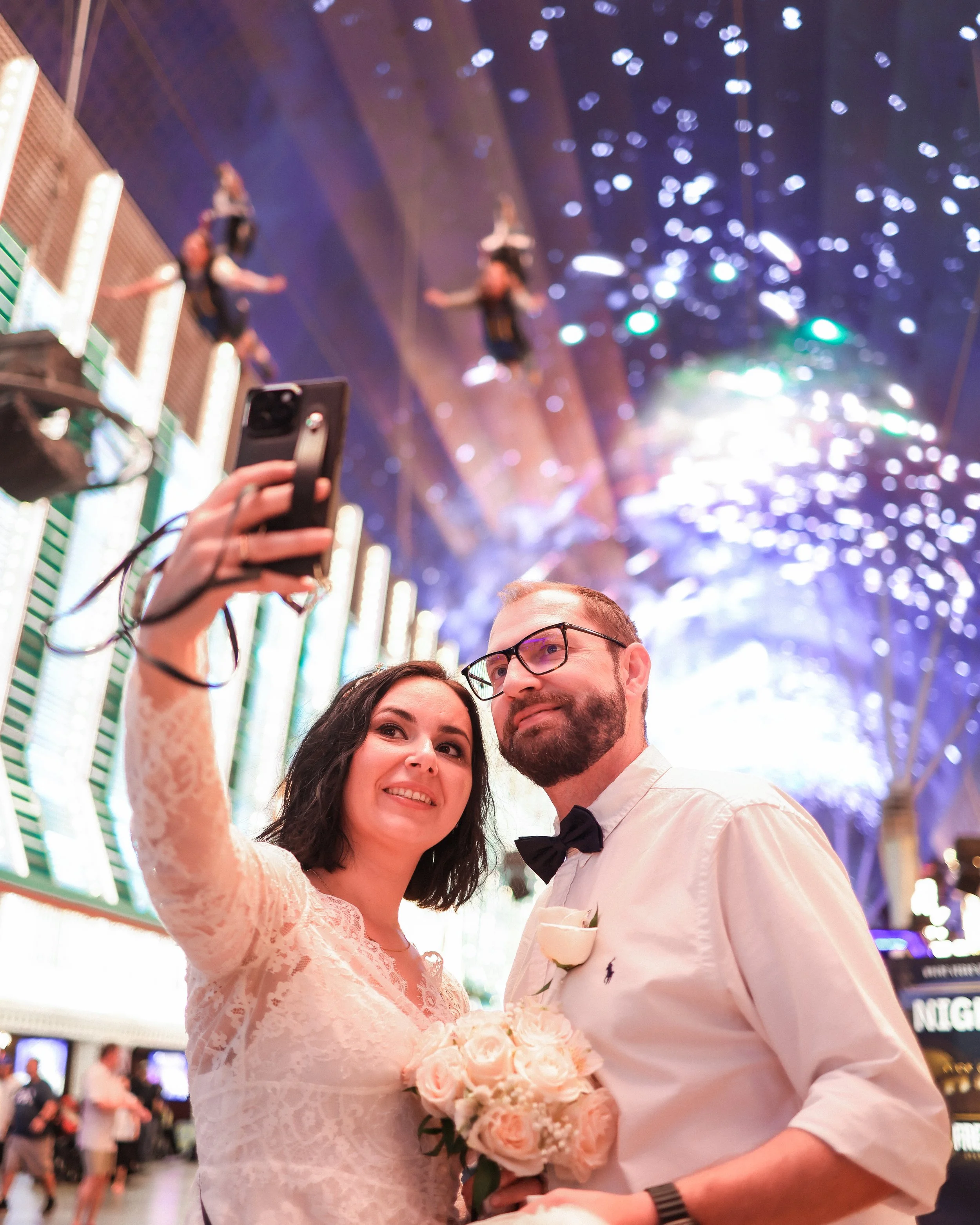 A smiling woman in a wedding dress holds a bouquet of white roses and takes a selfie with a man in tuxedo with a black bow tie, inside a festive venue with bright lights, colorful decorations, and other people in the background.