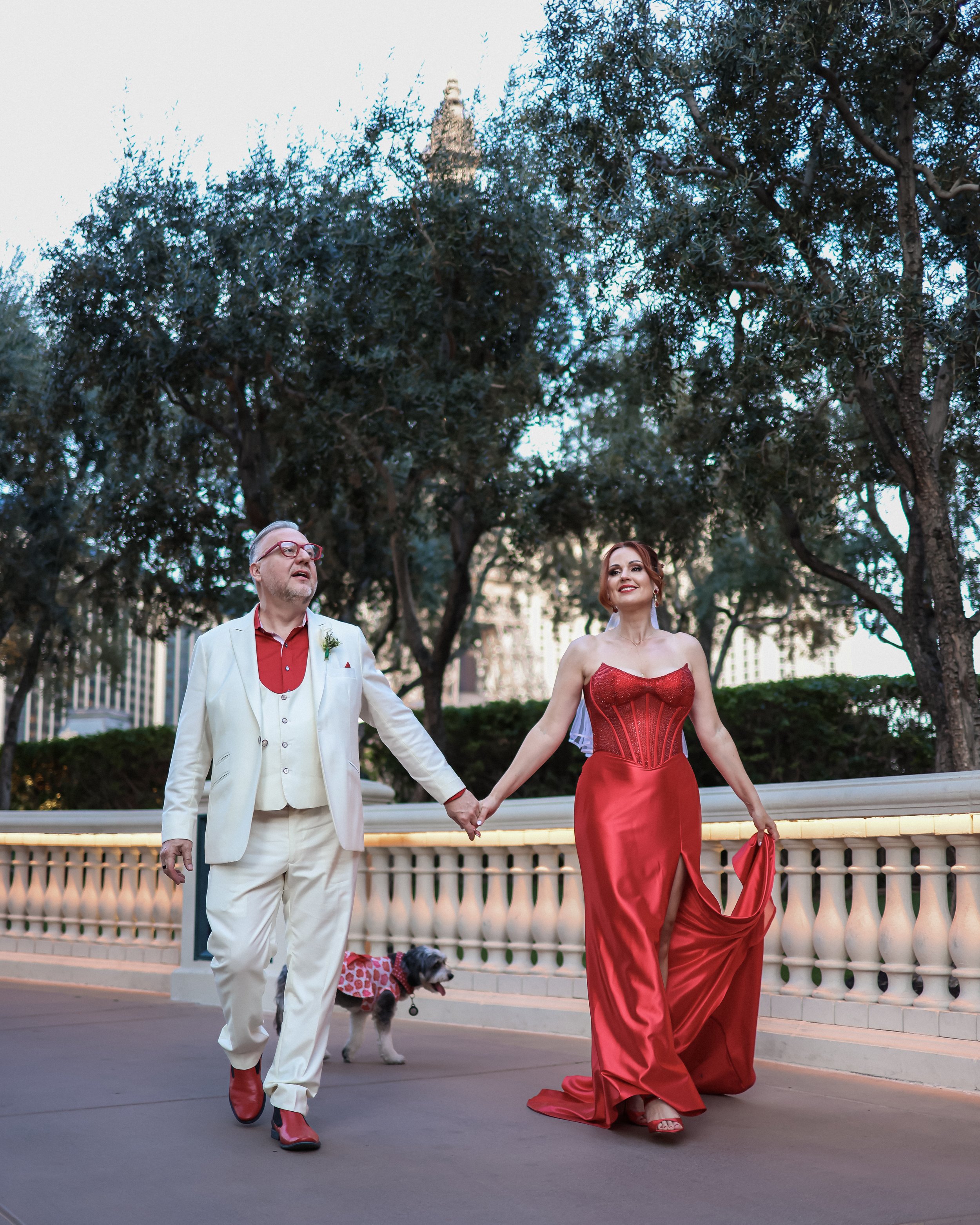 A couple dressed in formal attire walking hand in hand on a bridge in an outdoor setting, accompanied by a small dog in a pink polka dot outfit, with trees and city buildings in the background.