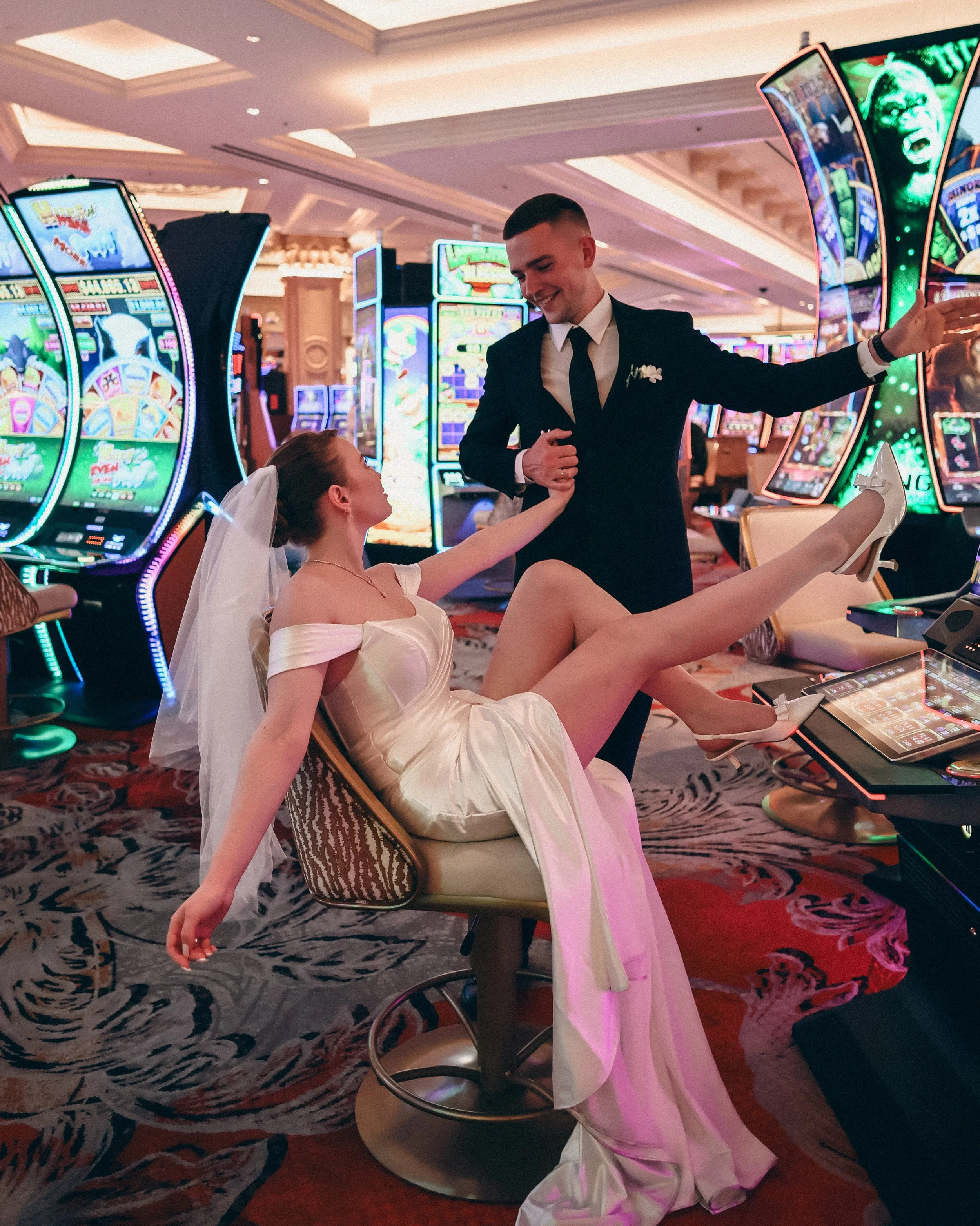 A wedding reception scene with a bride in a wedding dress and veil sitting on a chair, extending her leg with high heels on, and a groom in a tuxedo standing next to her, holding her hand, in front of colorful slot machines in a casino.