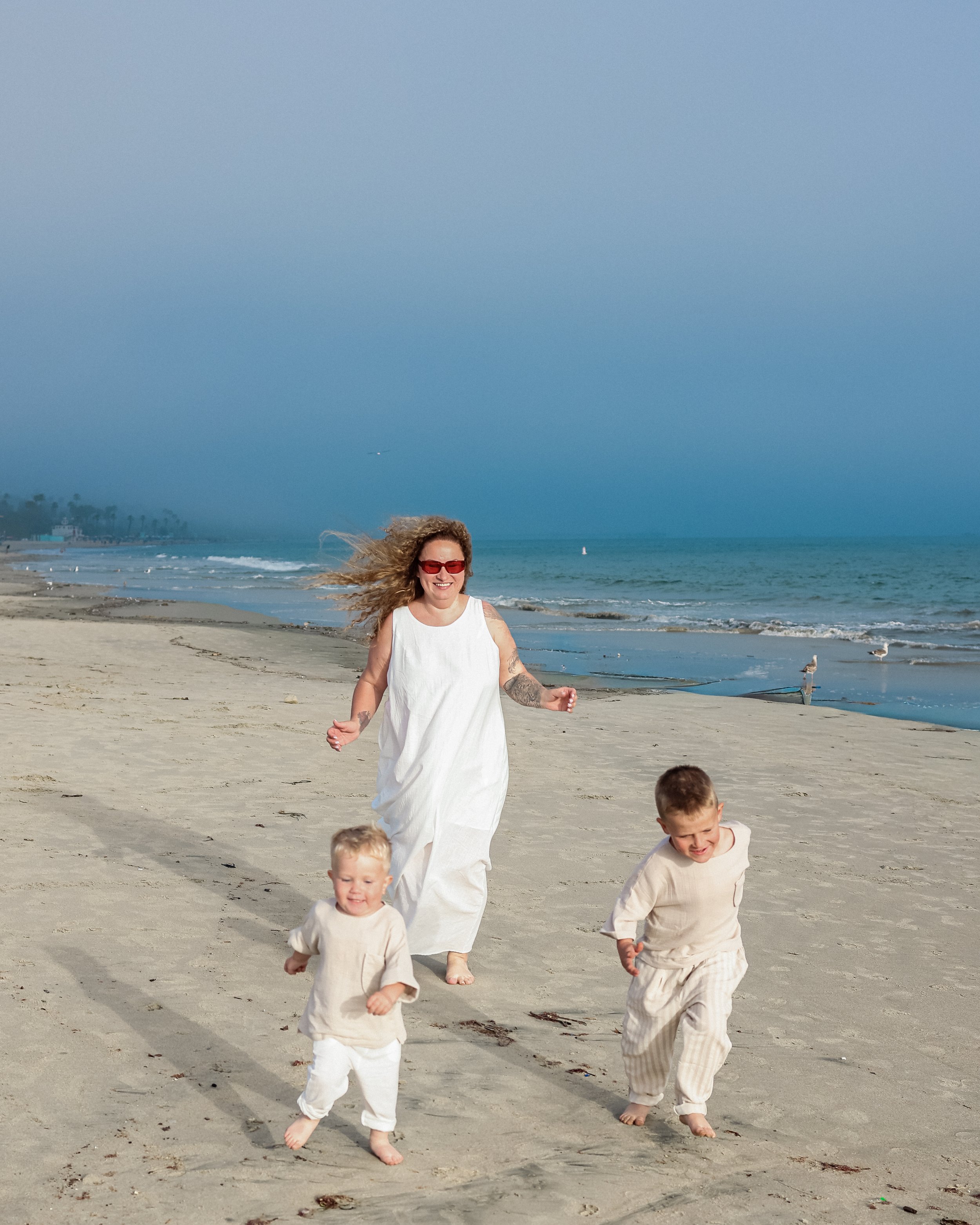 A woman with curly hair, foster sunglasses, and a white dress running on a beach with two young boys, one with blond hair and the other with darker hair, both in light-colored outfits, near the shoreline with the ocean and a cloudy sky in the background.