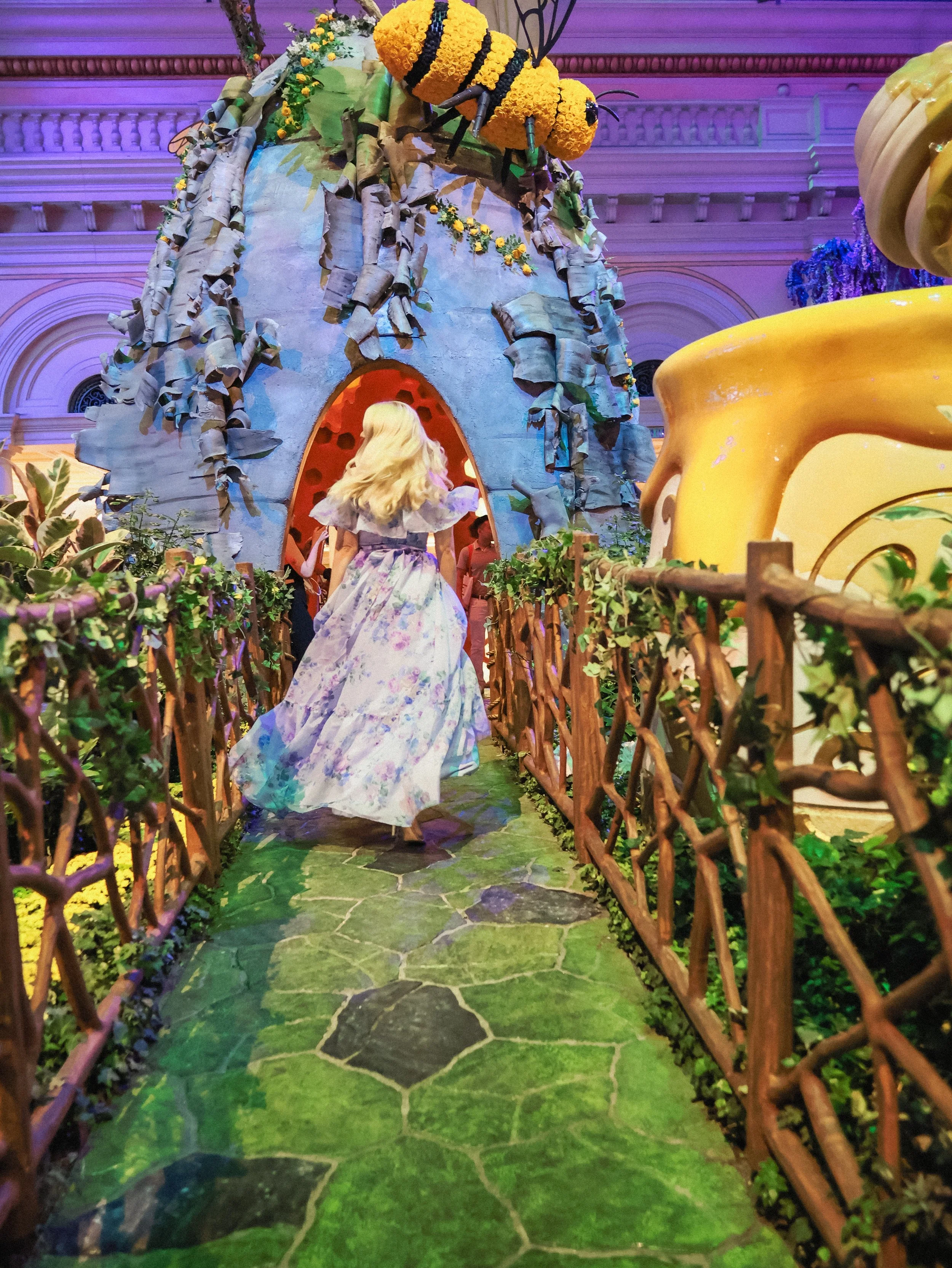 An indoor display resembling a whimsical fairy tale house with a large beehive decoration on top. A woman in a floral dress is walking along a path flanked by wooden fences and lush greenery.