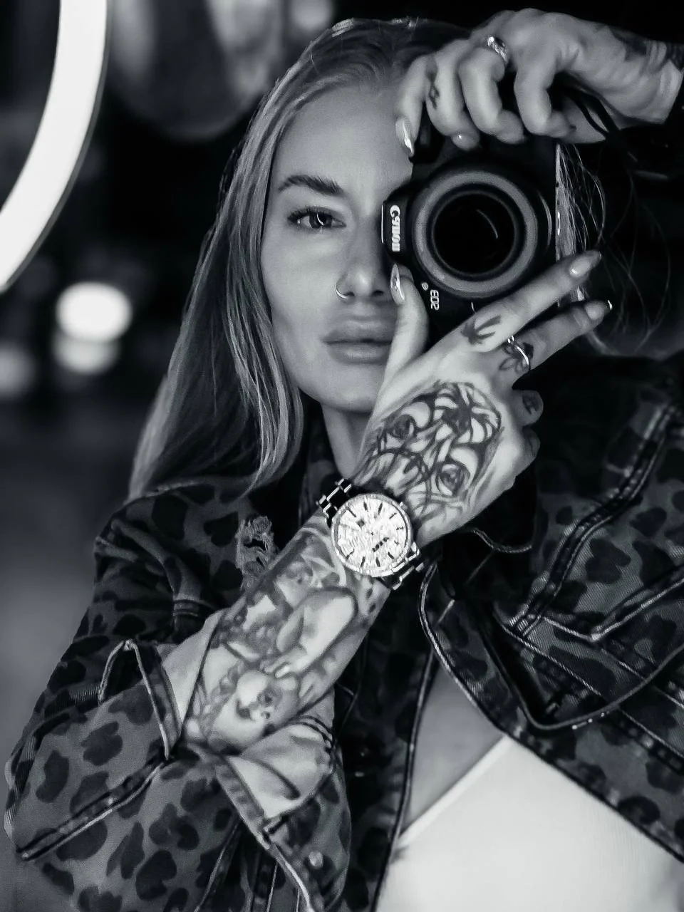 Black and white photo of a woman taking a mirror selfie with a camera, wearing a leopard-print jacket, with tattoos on her hands and wristwatch, and with long hair.