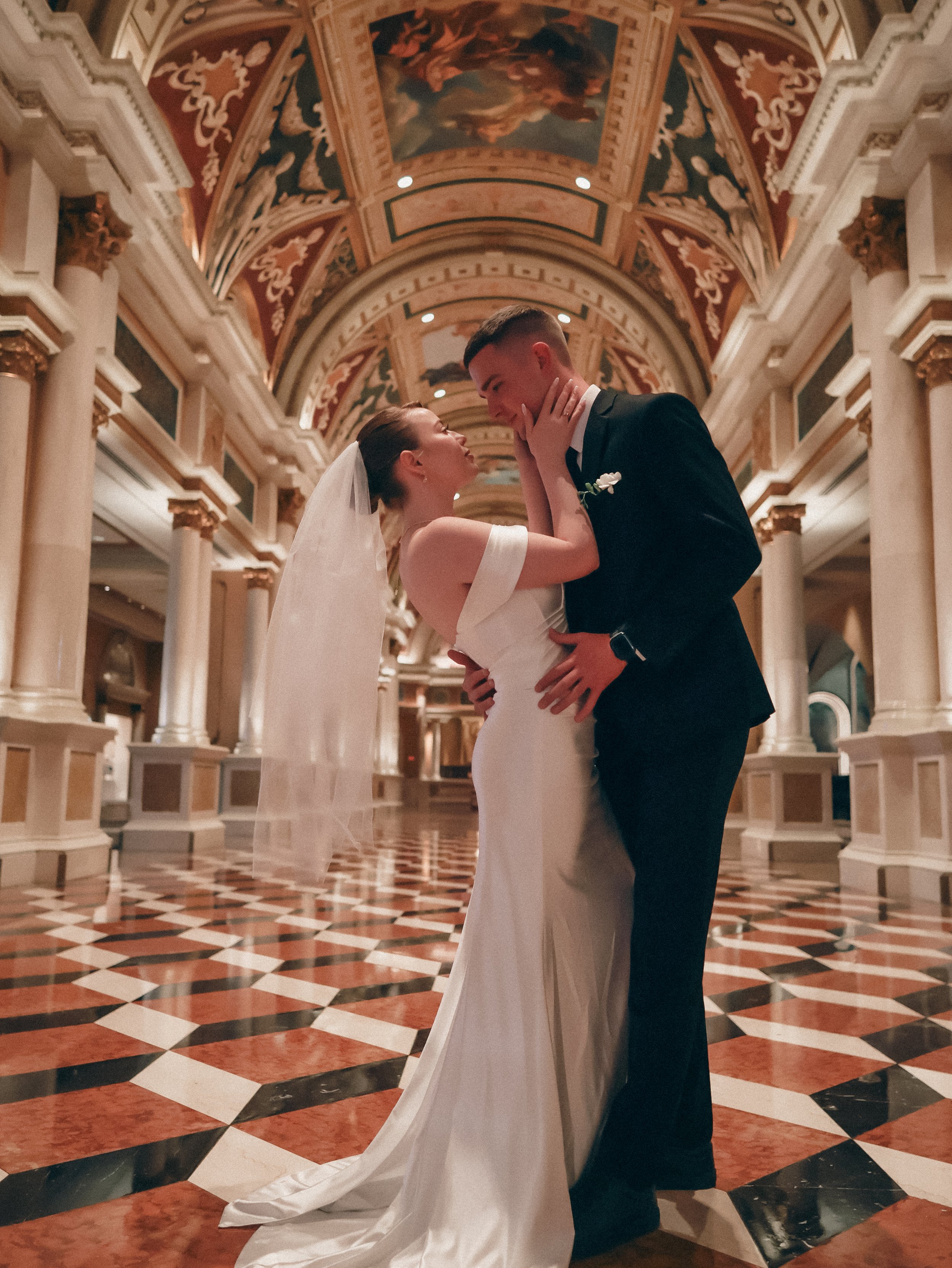 A bride and groom share a romantic moment in a grand hall with ornate ceiling artwork and black, white, and red checkered flooring.