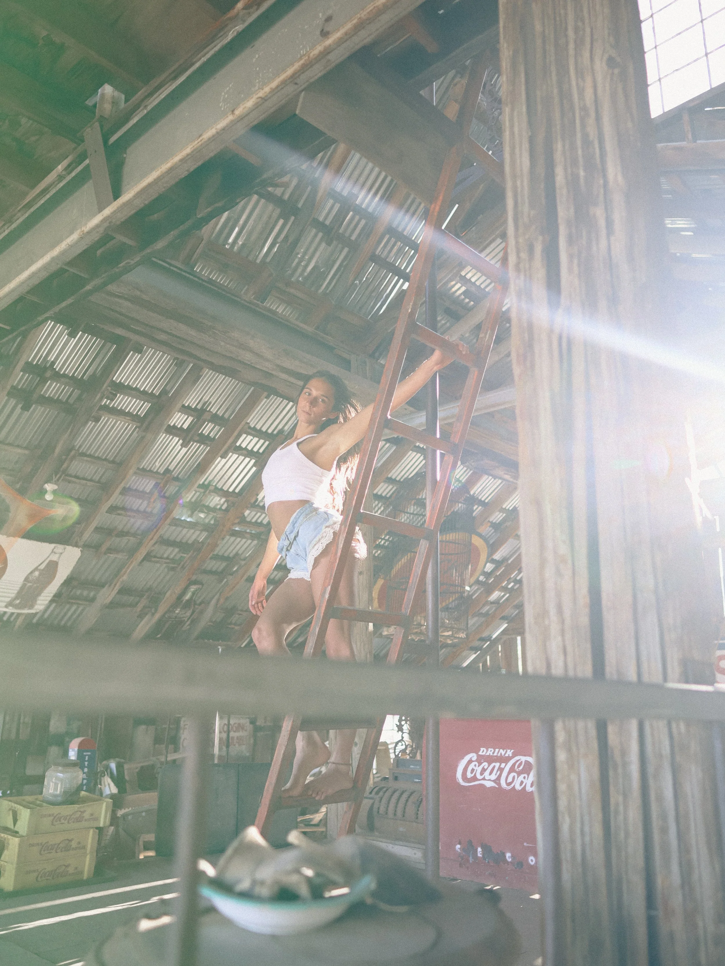 A person standing on a metal ladder inside a wooden building, reaching up towards the roof. The scene is illuminated by sunlight streaming in through gaps in the roof. The foreground shows a counter with a small bowl, a Coca-Cola vending machine, and