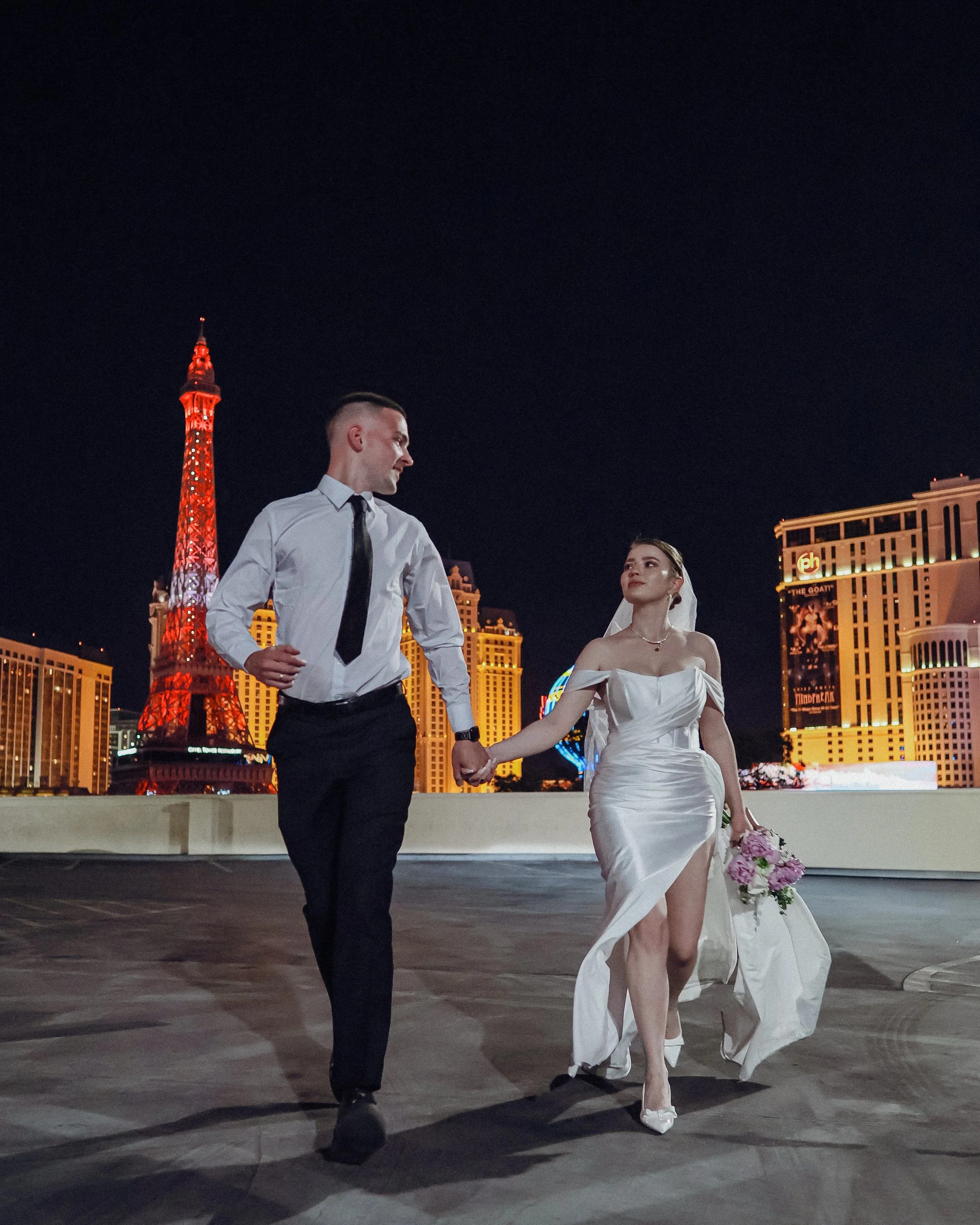 A man and woman in formal attire holding hands, walking together at night with the Las Vegas Strip and the Eiffel Tower replica illuminated in the background.