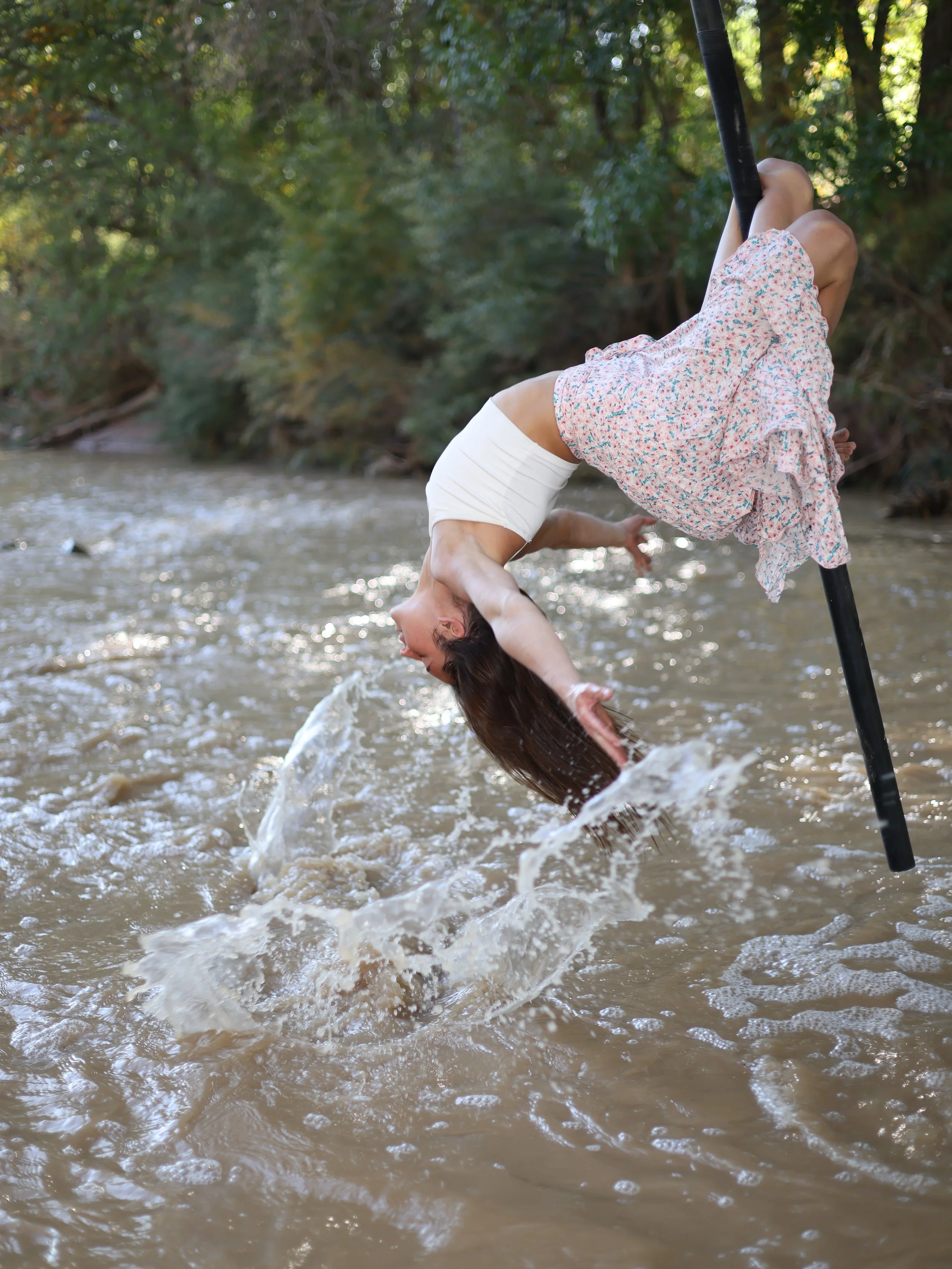 A woman in a floral dress and white shorts is performing a backflip over a shallow river with a forested background.