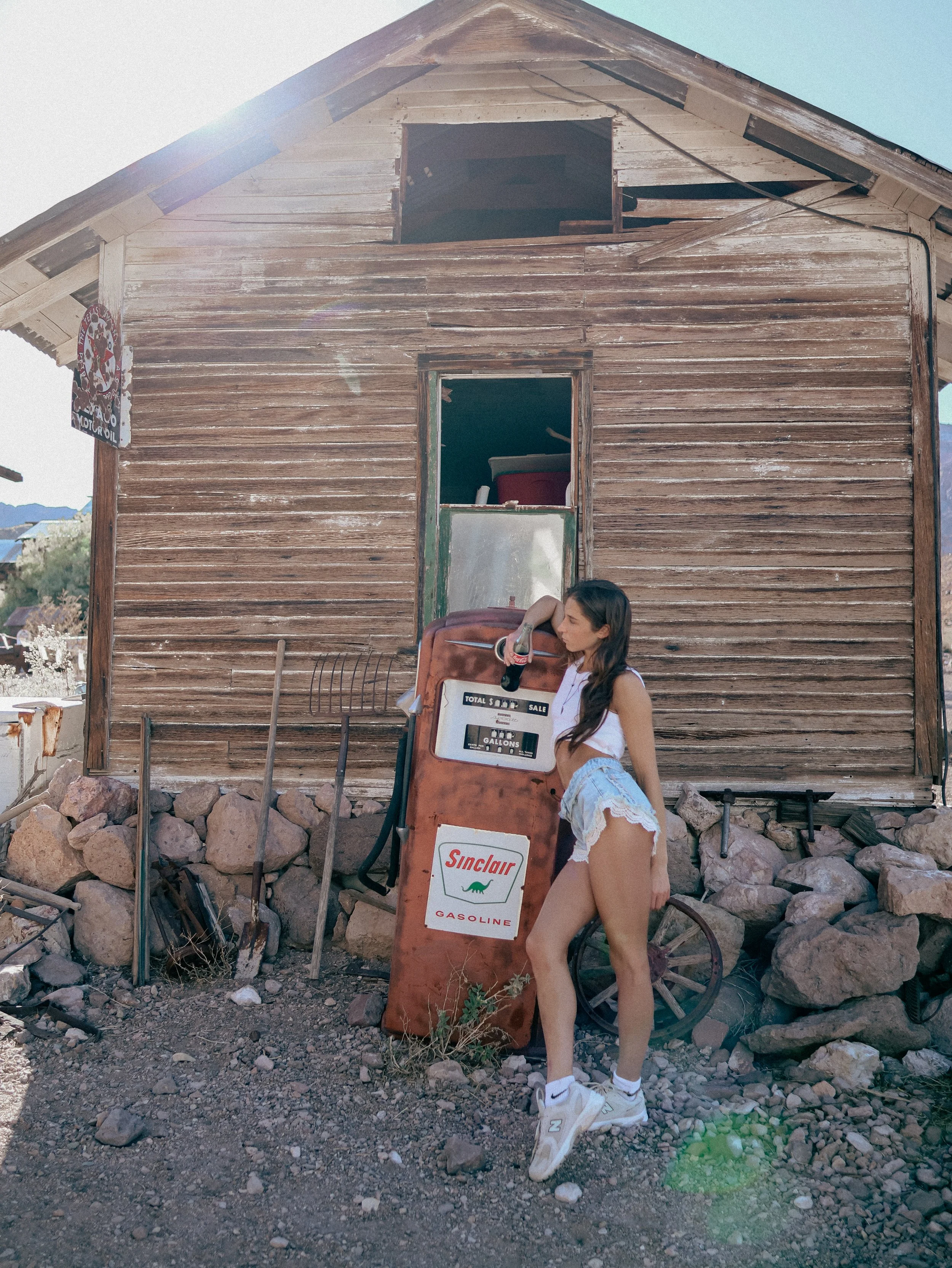 A young woman with long dark hair, wearing a white tank top, patterned shorts, and white sneakers, is standing outdoors in front of an old wooden building with weathered siding. She is leaning against or resting her arm on a vintage gas pump that has