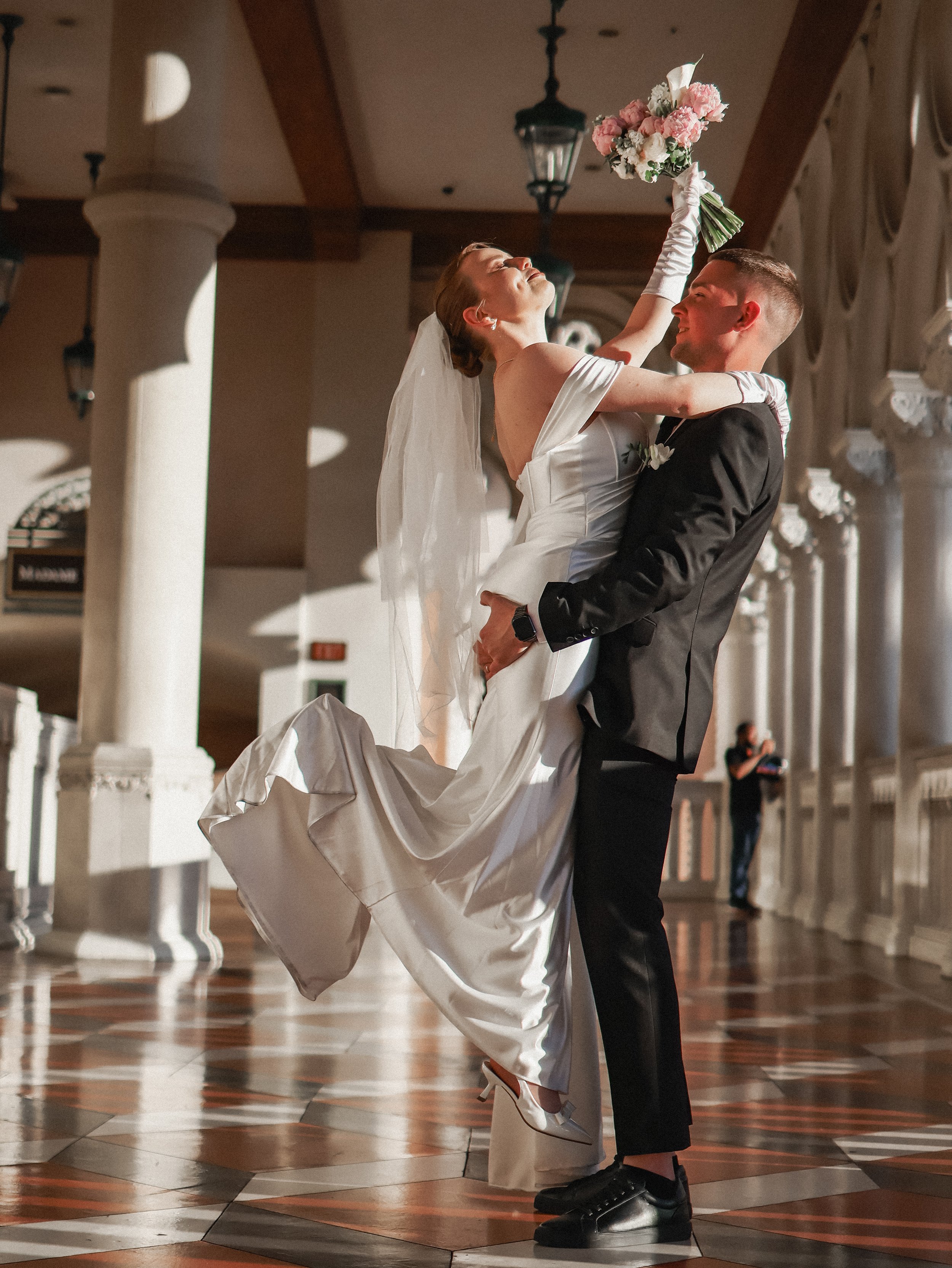 A bride is lifted by a groom, holding a bouquet of flowers, inside a decorated indoor venue with large pillars and patterned tile floor.