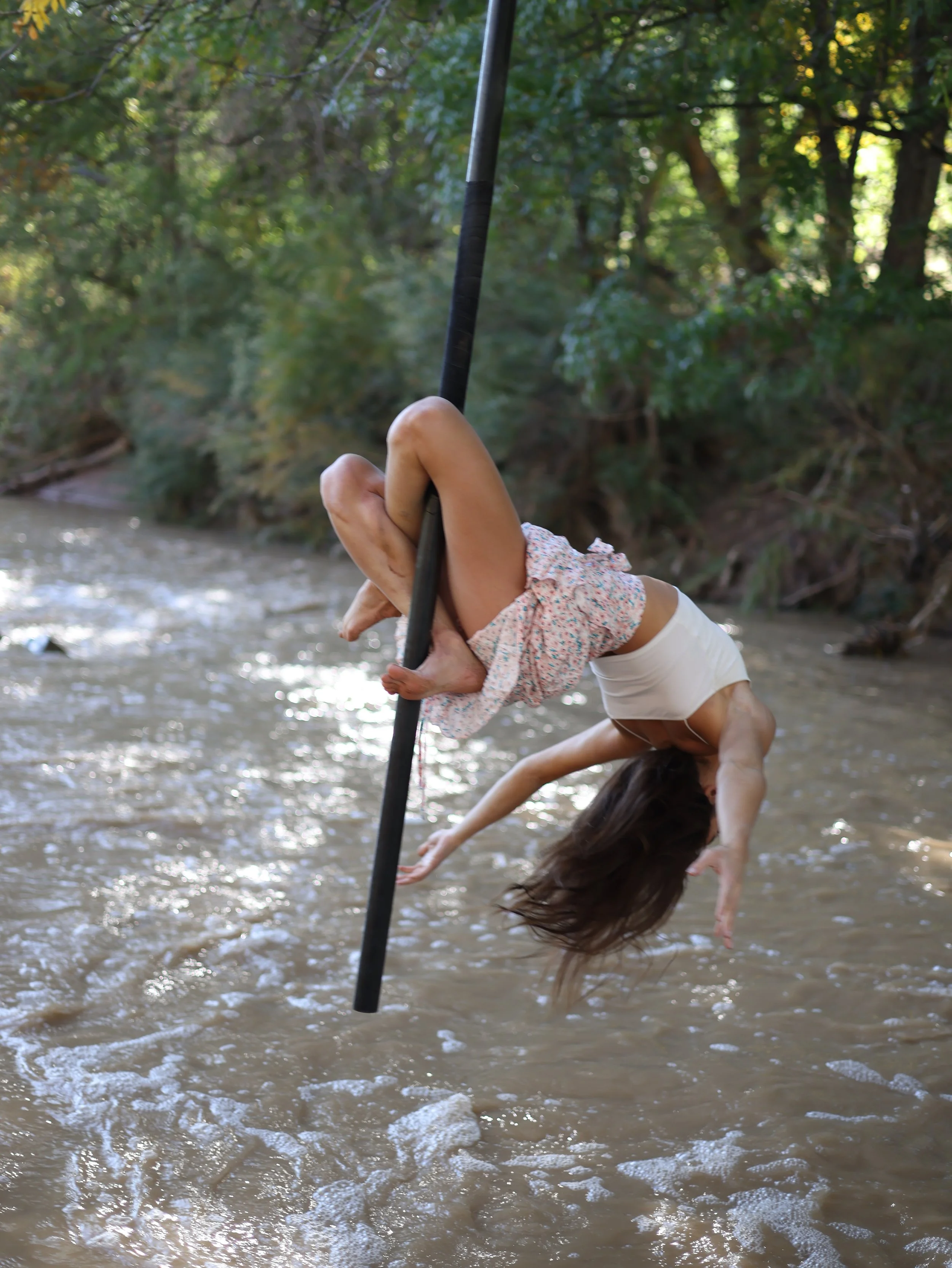 A woman performing a handstand on a pole in a shallow river in a forested area with trees and water.