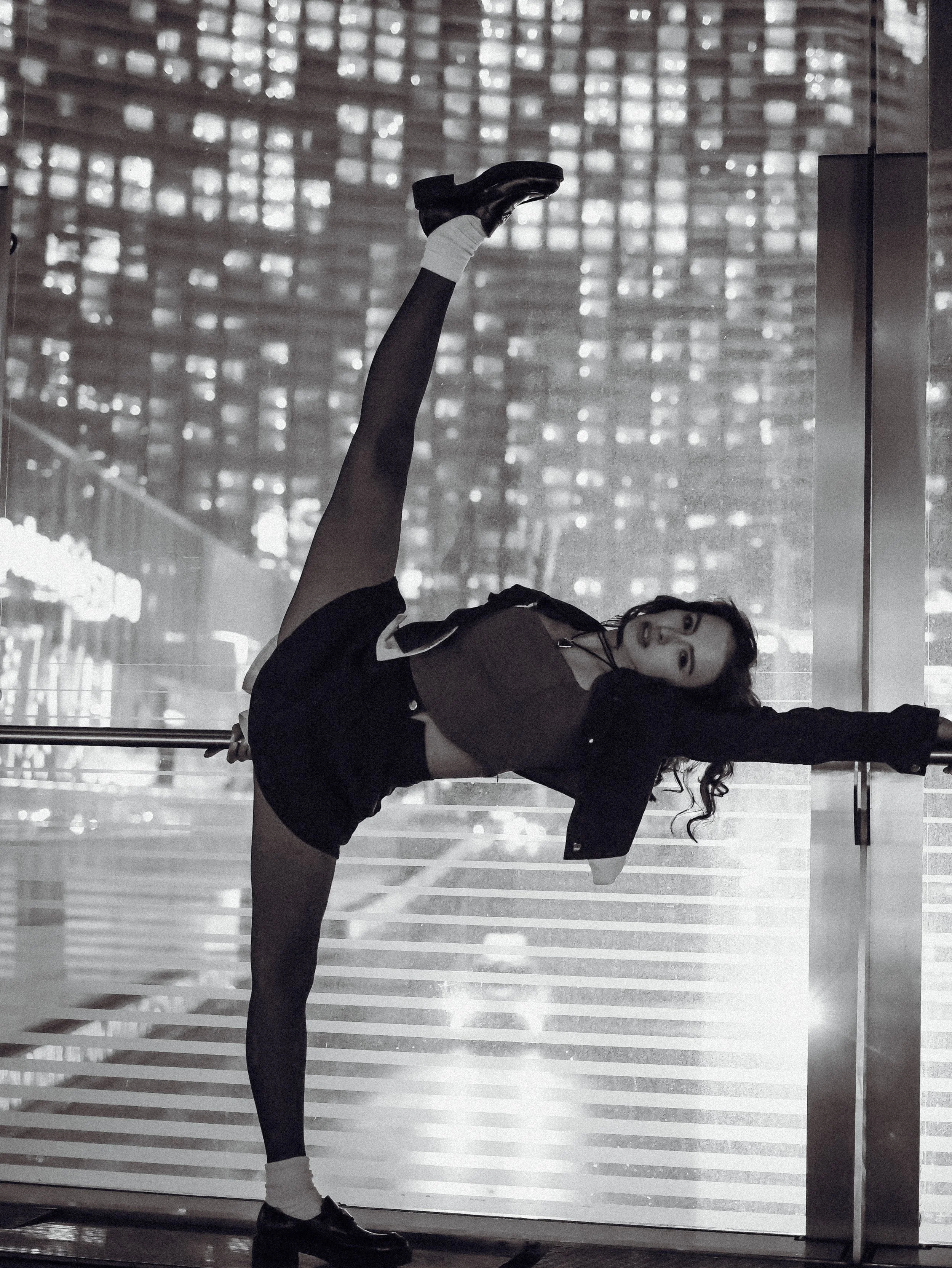 A woman with curly hair, wearing a dark jacket, shorts, and sneakers, is standing in front of a glass window with a cityscape background. She is holding her ankle in a stretch.
