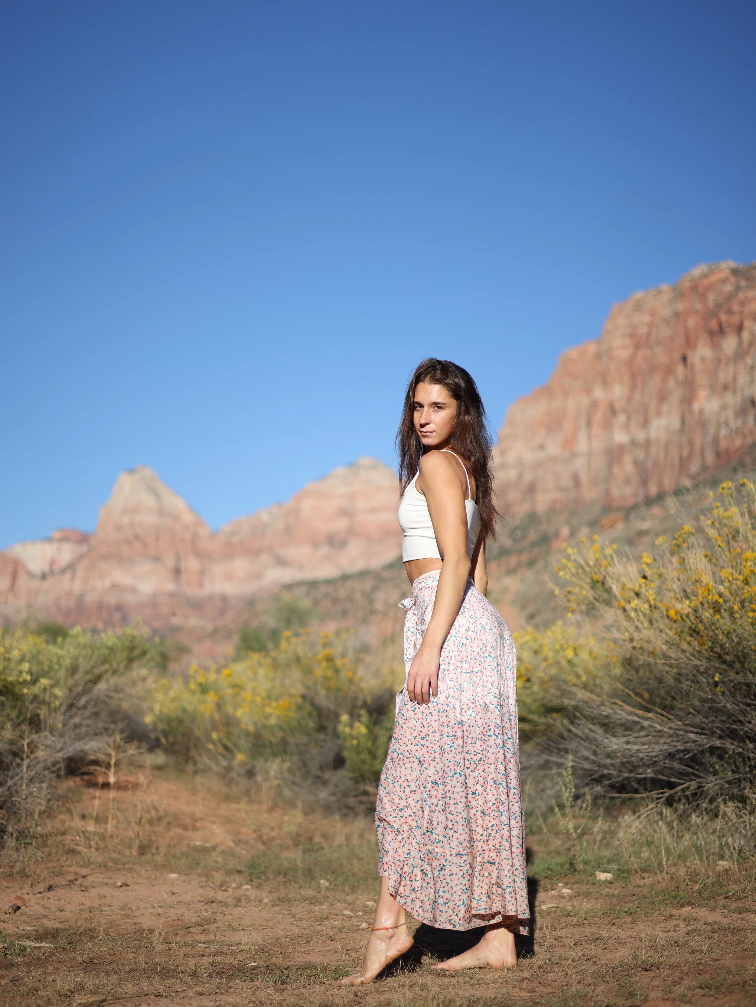 A young woman standing barefoot in a desert landscape with rocky formations in the background, wearing a sleeveless white top and a long, flowing patterned skirt, with the sun casting shadows.