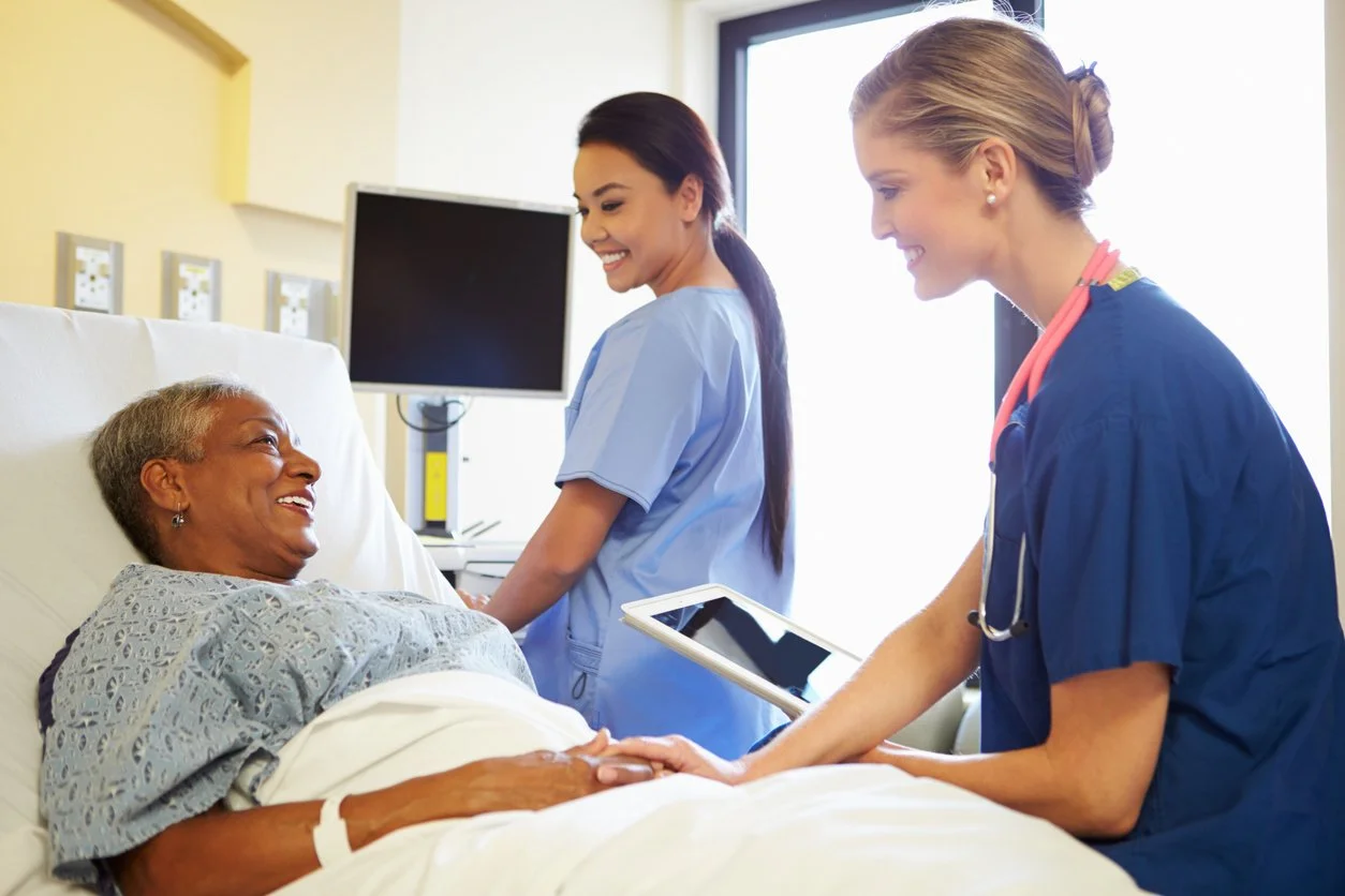 Two healthcare professionals, one female nurse and one female doctor, smiling and holding hands with an elderly female patient lying in a hospital bed, in a well-lit hospital room.