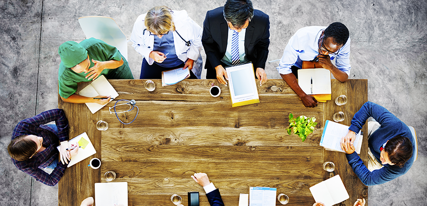 Top-down view of a diverse group of people sitting around a large wooden conference table, engaging with various documents, laptops, and notebooks, with a small plant and glasses of water on the table.