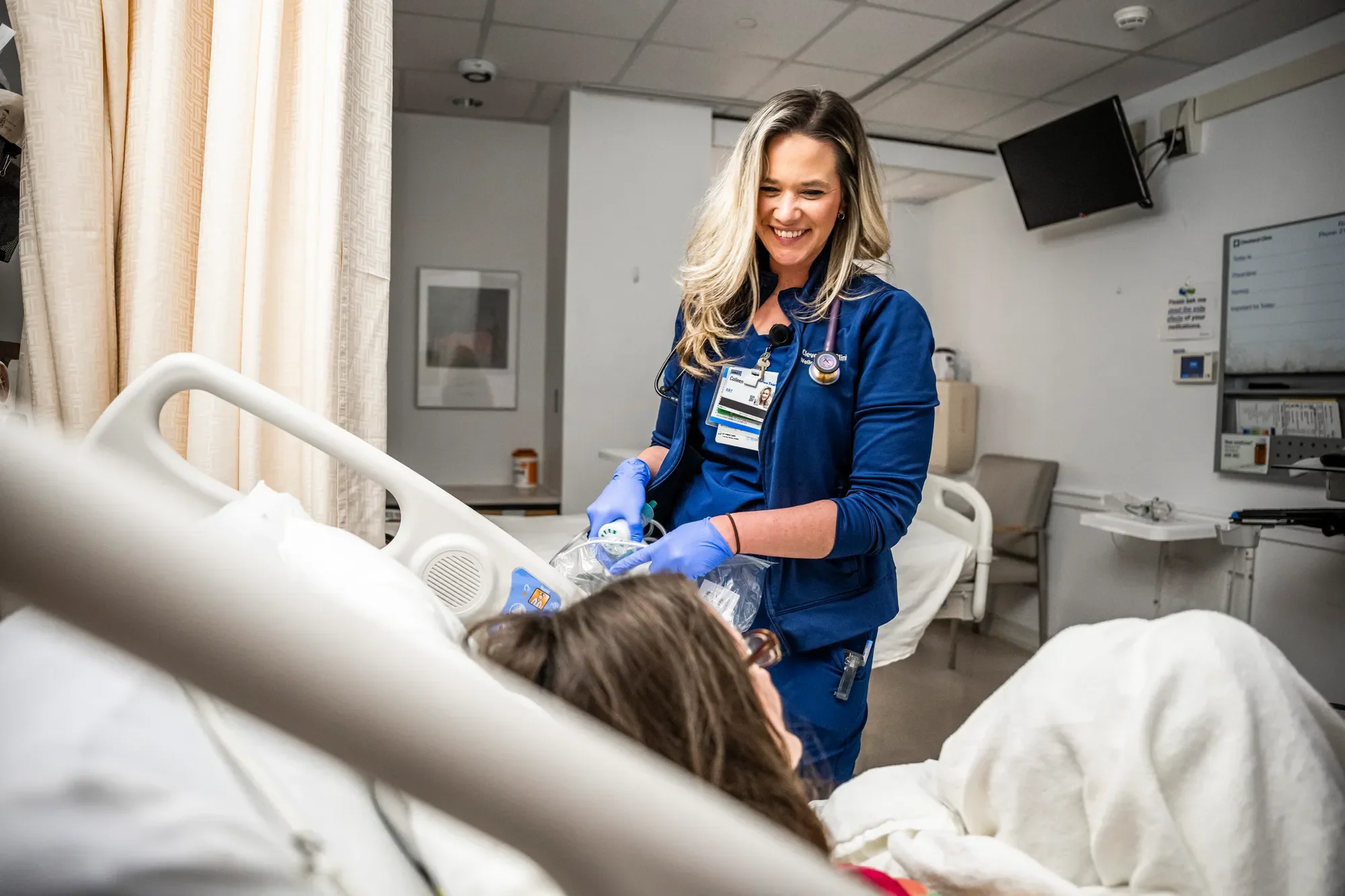 A smiling female healthcare worker in blue scrubs and gloves helps a patient in a hospital bed.