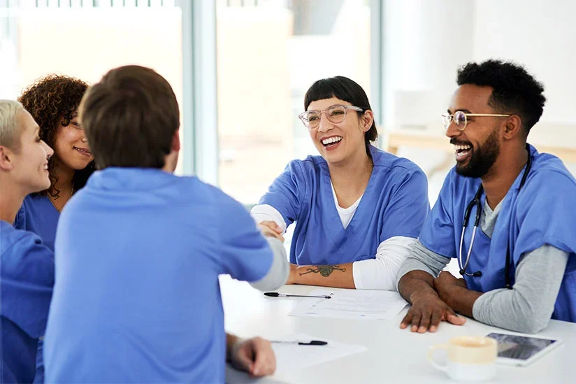 A diverse group of healthcare professionals, including women and men of different ethnicities, in blue scrubs, smiling and shaking hands during a meeting in a bright, modern office.