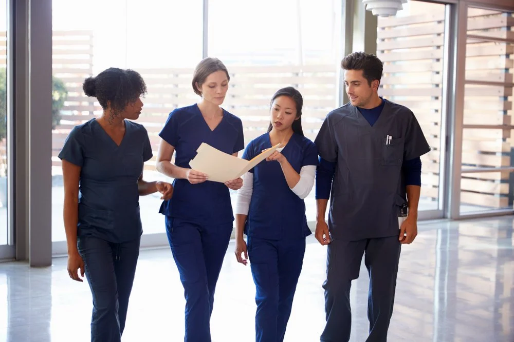 Four healthcare professionals, two women and two men, walking together in a hospital or clinic. They are wearing navy blue scrubs and appear to be discussing patient records or a report.