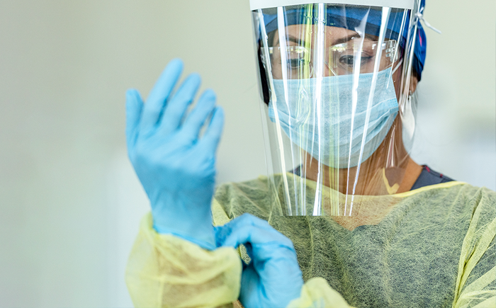 Healthcare worker wearing a face shield, safety mask, blue gloves, and yellow protective gown, preparing to put on gloves.