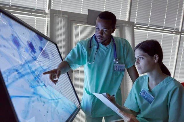 Two healthcare professionals, a male and a female nurse, analyze medical images on a digital touchscreen monitor in a hospital setting.