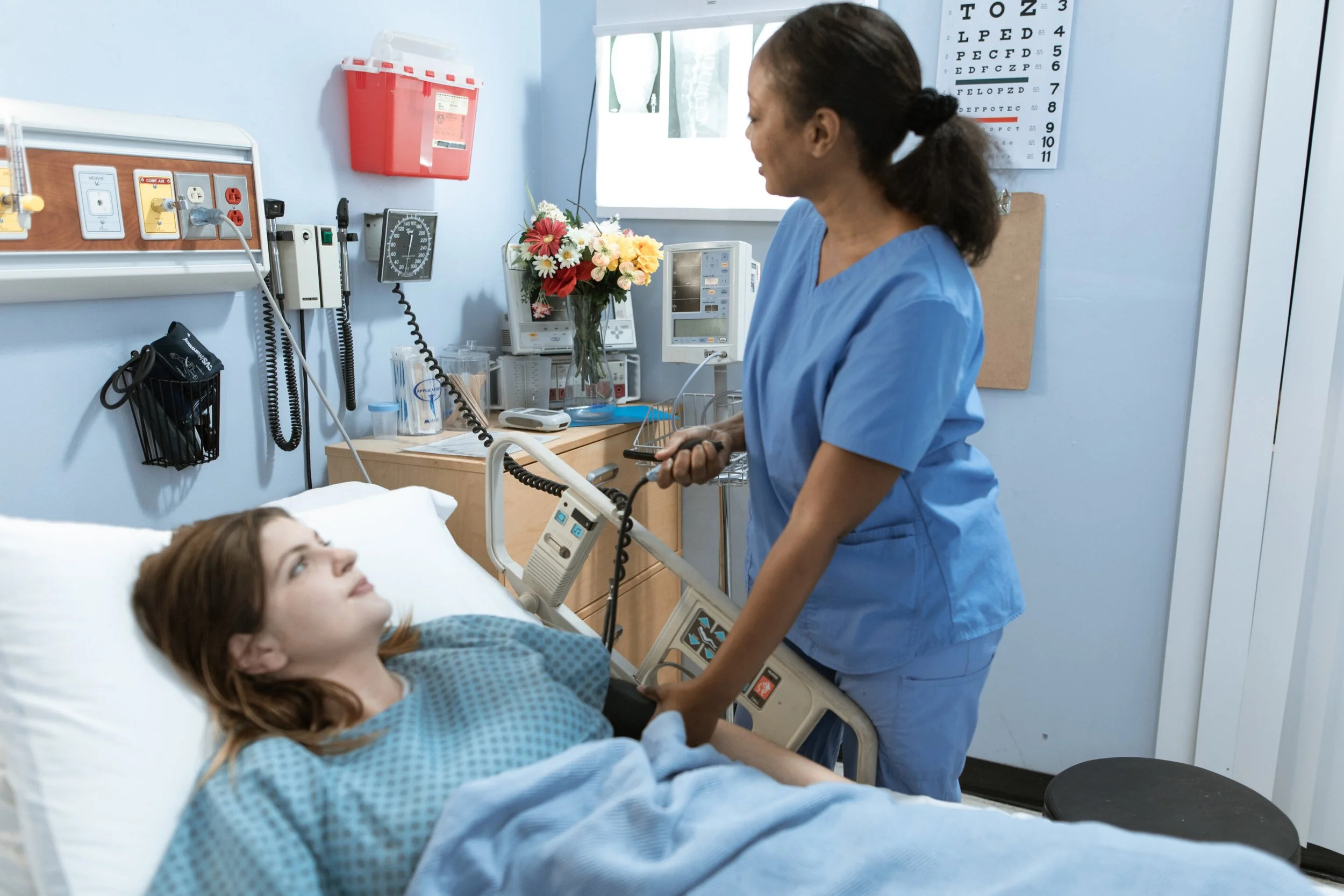 A nurse in blue scrubs holds a patient's hand and checks her vital signs in a hospital room.
