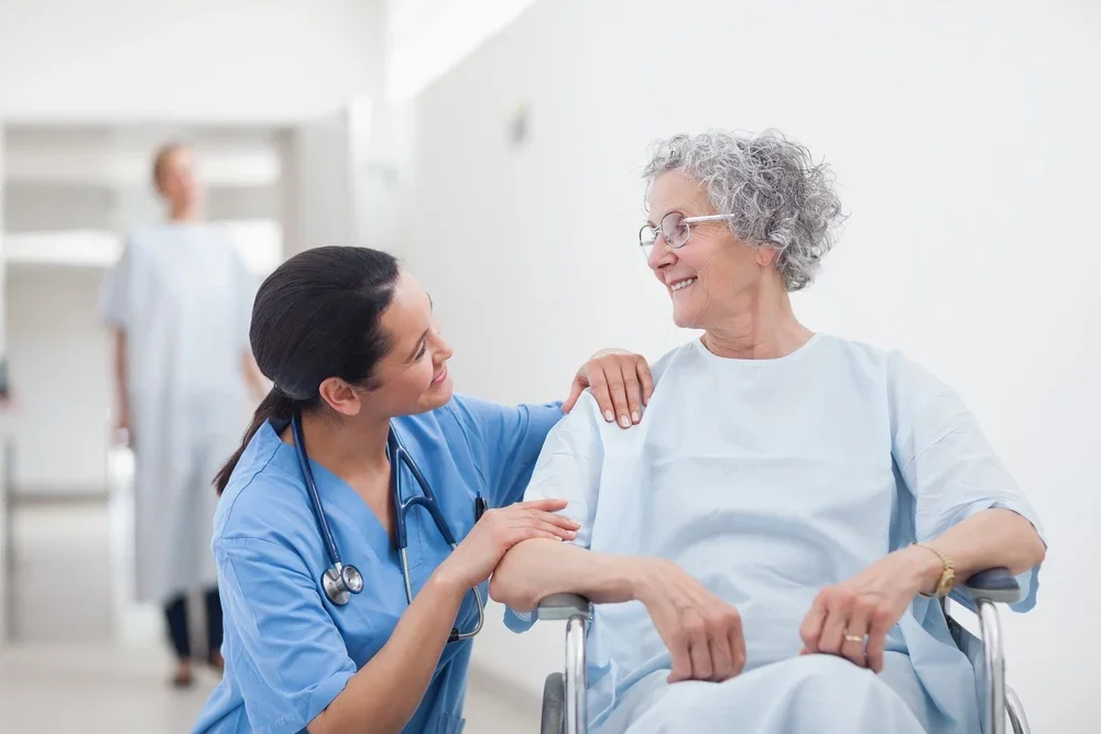 A healthcare professional, wearing scrubs and a stethoscope, talking to an elderly woman in a hospital gown who is sitting in a wheelchair, both smiling. A person in medical attire is blurred in the background.
