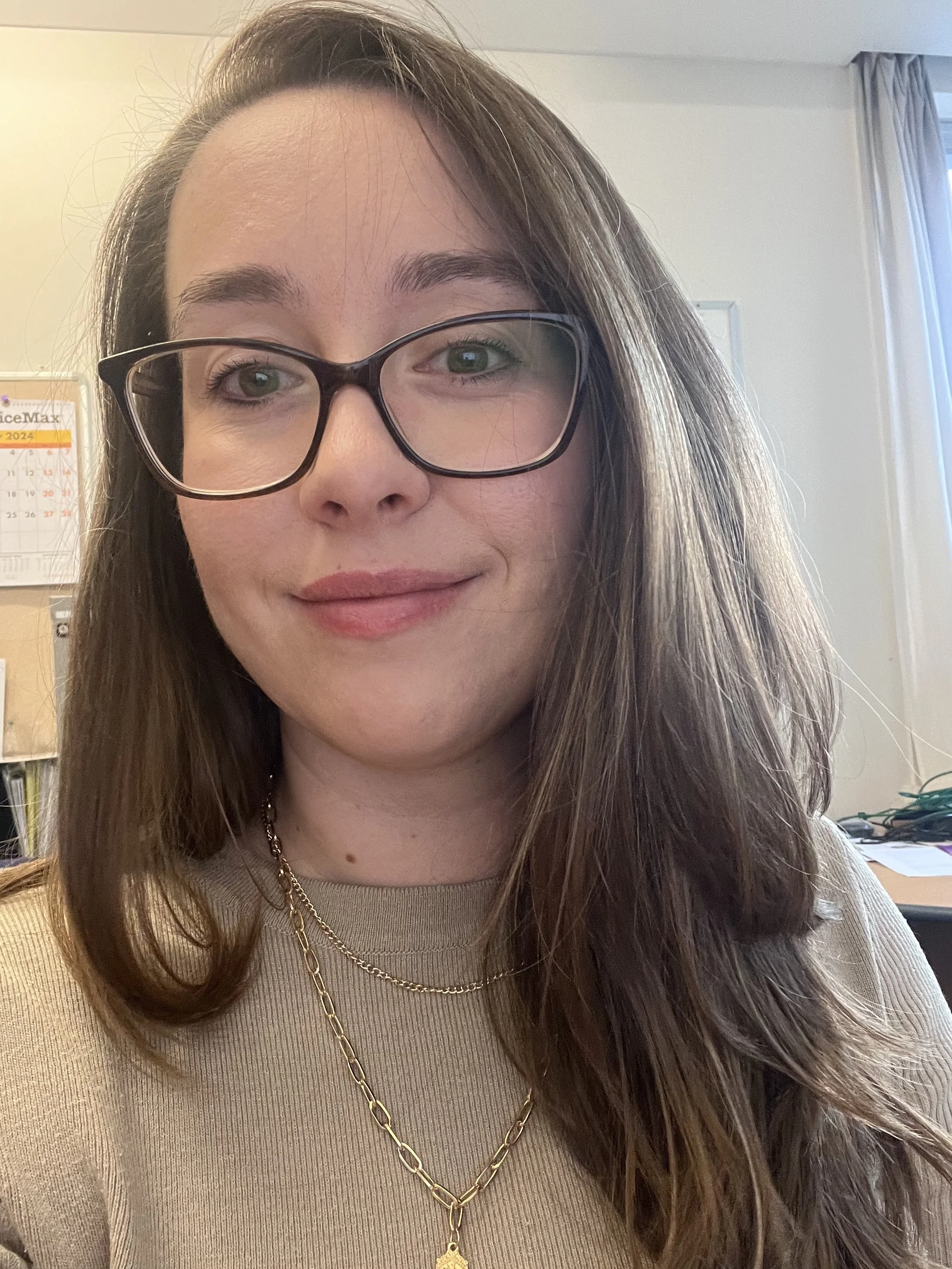 A woman with long brown hair, glasses, and light makeup smiling indoors, wearing a beige top and gold necklaces, with a calendar and office items in the background.