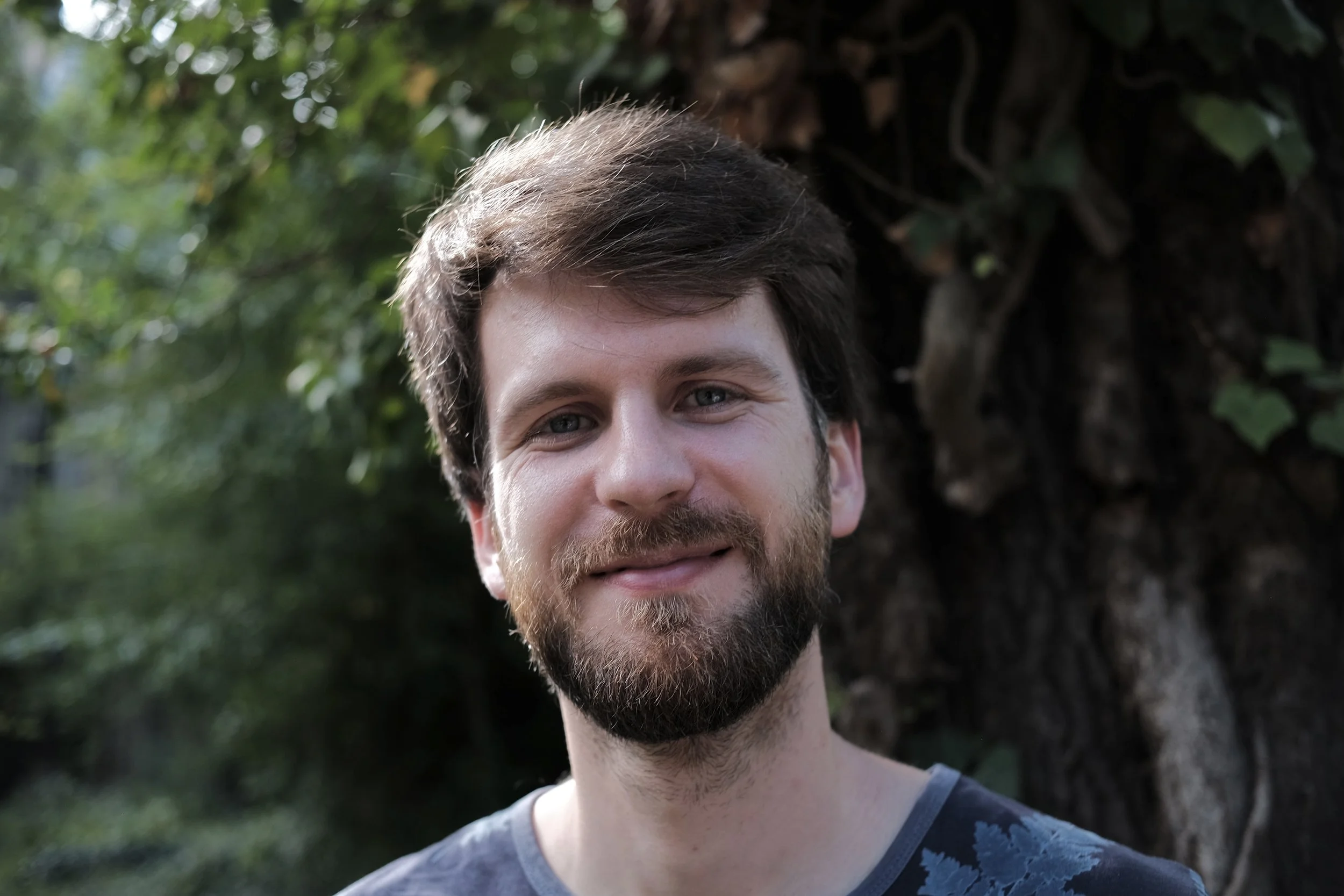 A smiling young man with light skin, brown hair, and a beard, standing outdoors near a large tree with lush green leaves.