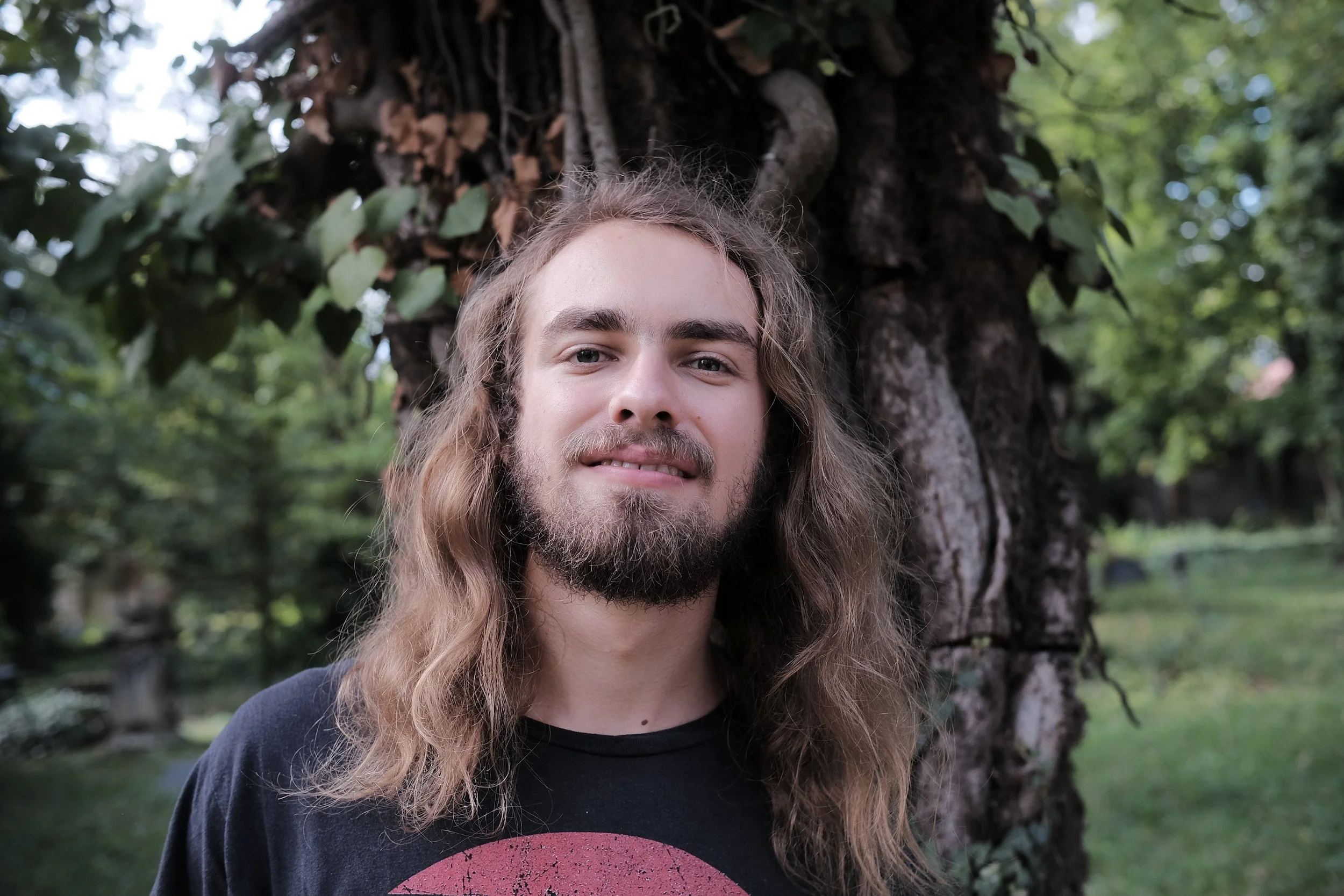 A man with long hair and a beard stands outdoors in front of a tree with green leaves.