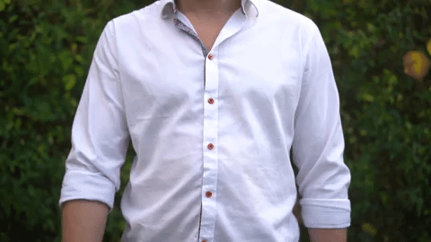 Person wearing a white button-up shirt with rolled sleeves standing outdoors in front of green foliage.